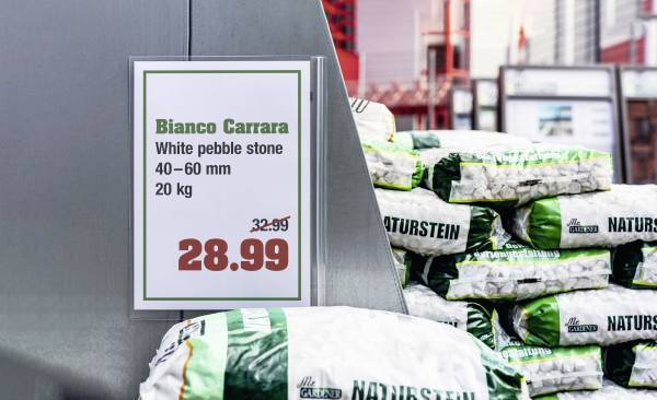 Ein Preisschild zeigt 'Bianco Carrara', weiße Kieselsteine, 40-60 mm, 20 kg, für 28,99 EUR. Es gibt gestapelte Säcke im Hintergrund.