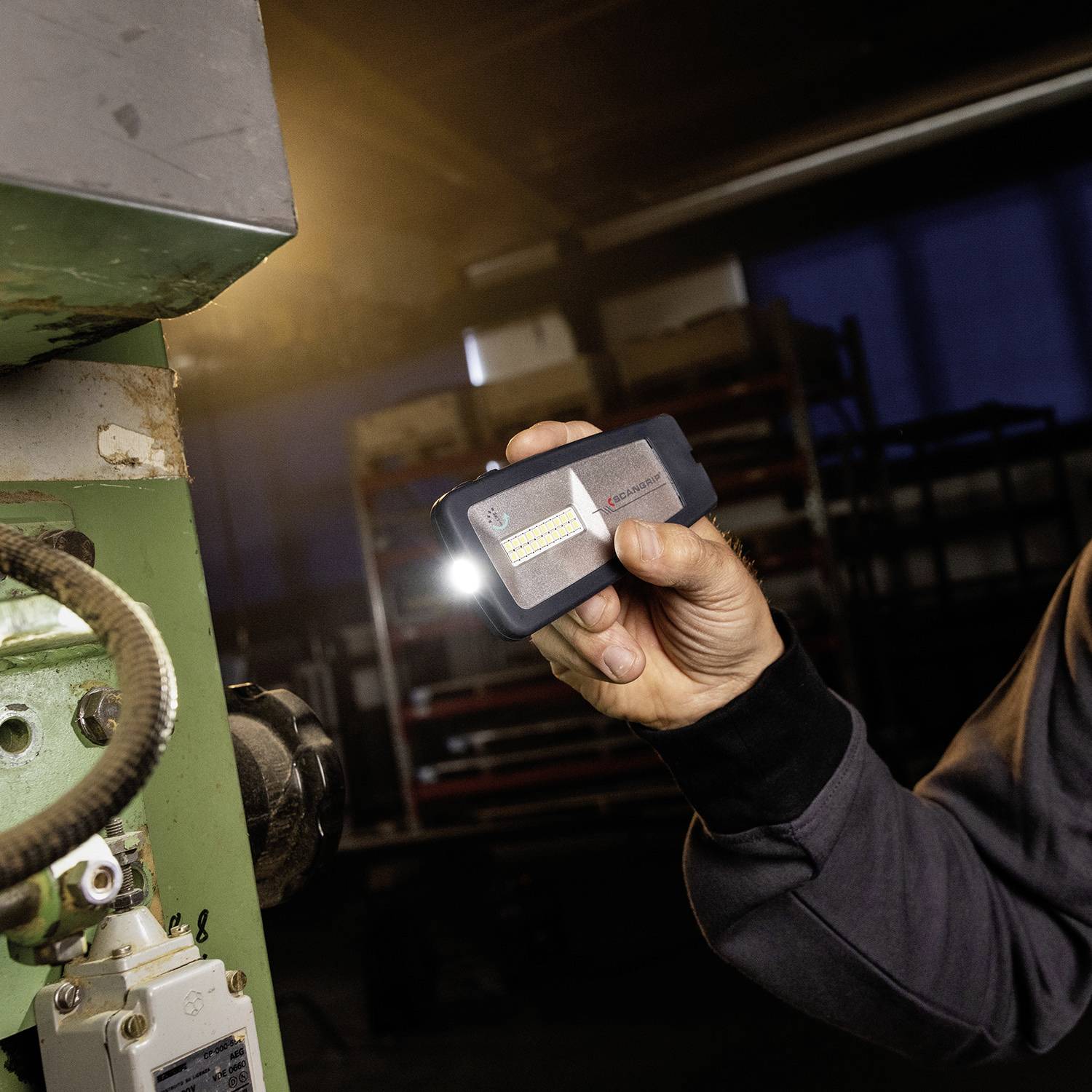 Eine Person hält ein kleines, rechteckiges Messgerät mit einem leuchtenden Licht in der Hand. Die Umgebung wirkt industriell.