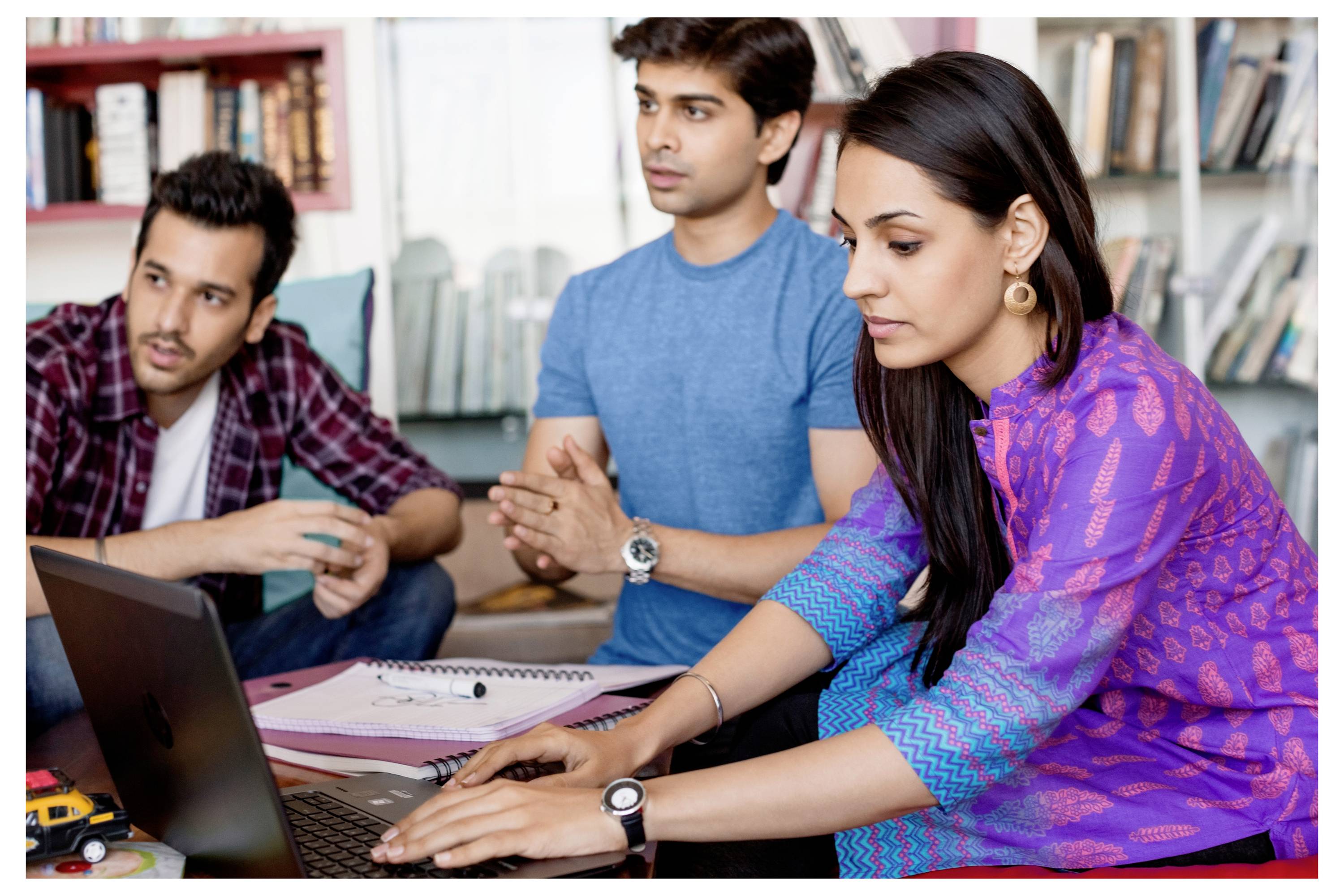Drei Personen arbeiten an einem Laptop in einer Bibliothek. Eine Frau tippt, während zwei Männer aufmerksam zuschauen. Bücherregale im Hintergrund.