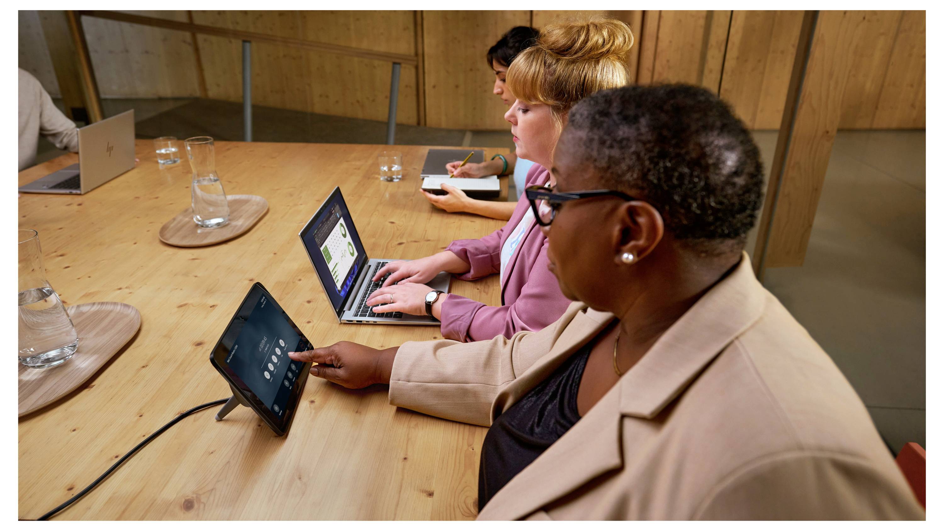 Drei Personen an einem Holztisch mit Laptops, eine Person tippt auf ein Tablet. Besprechung oder Arbeitssituation in modernem Büro.