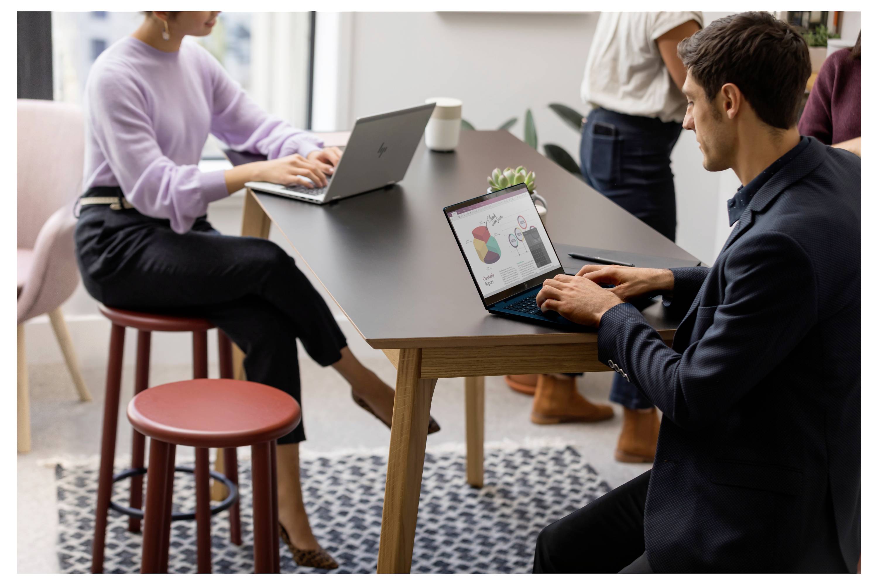 Eine Gruppe von Menschen arbeitet in einem Büroraum. Zwei Personen sitzen an einem Tisch und verwenden Laptops. Ein Diagramm ist auf einem Bildschirm sichtbar.
