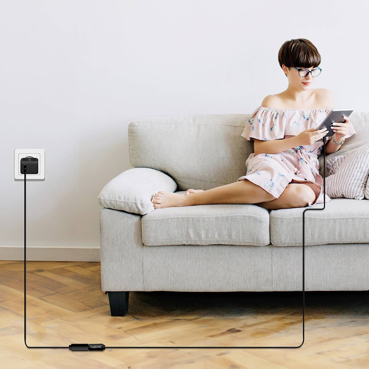 Person mit Tablet sitzt auf einem Sofa. Ein angeschlossenes Ladekabel führt zur Steckdose. Raum hat Holzboden und weiße Wände.