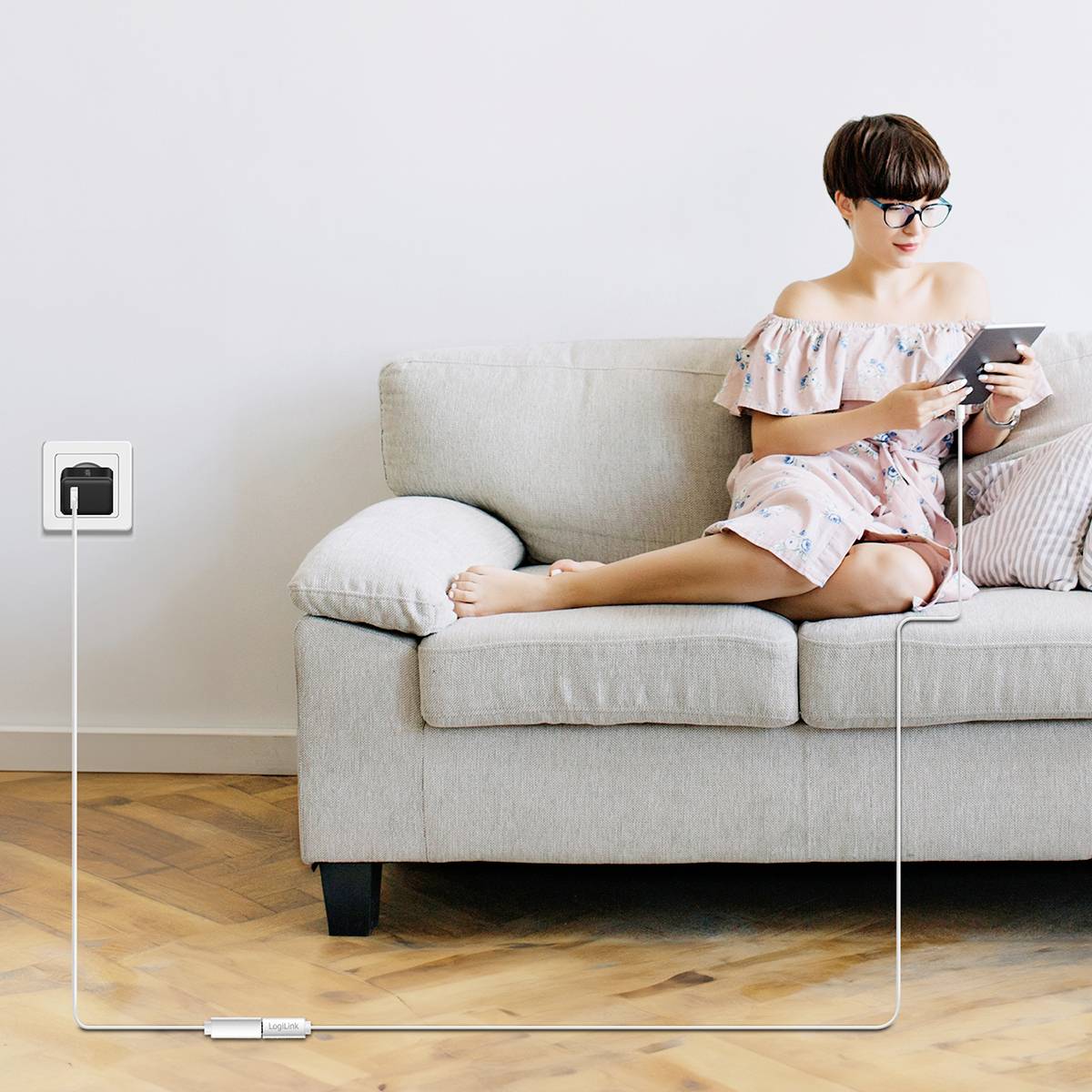 Eine Person sitzt auf einem Sofa und verwendet ein Smartphone. Ein Ladekabel ist mit einer Steckdose an der Wand verbunden.