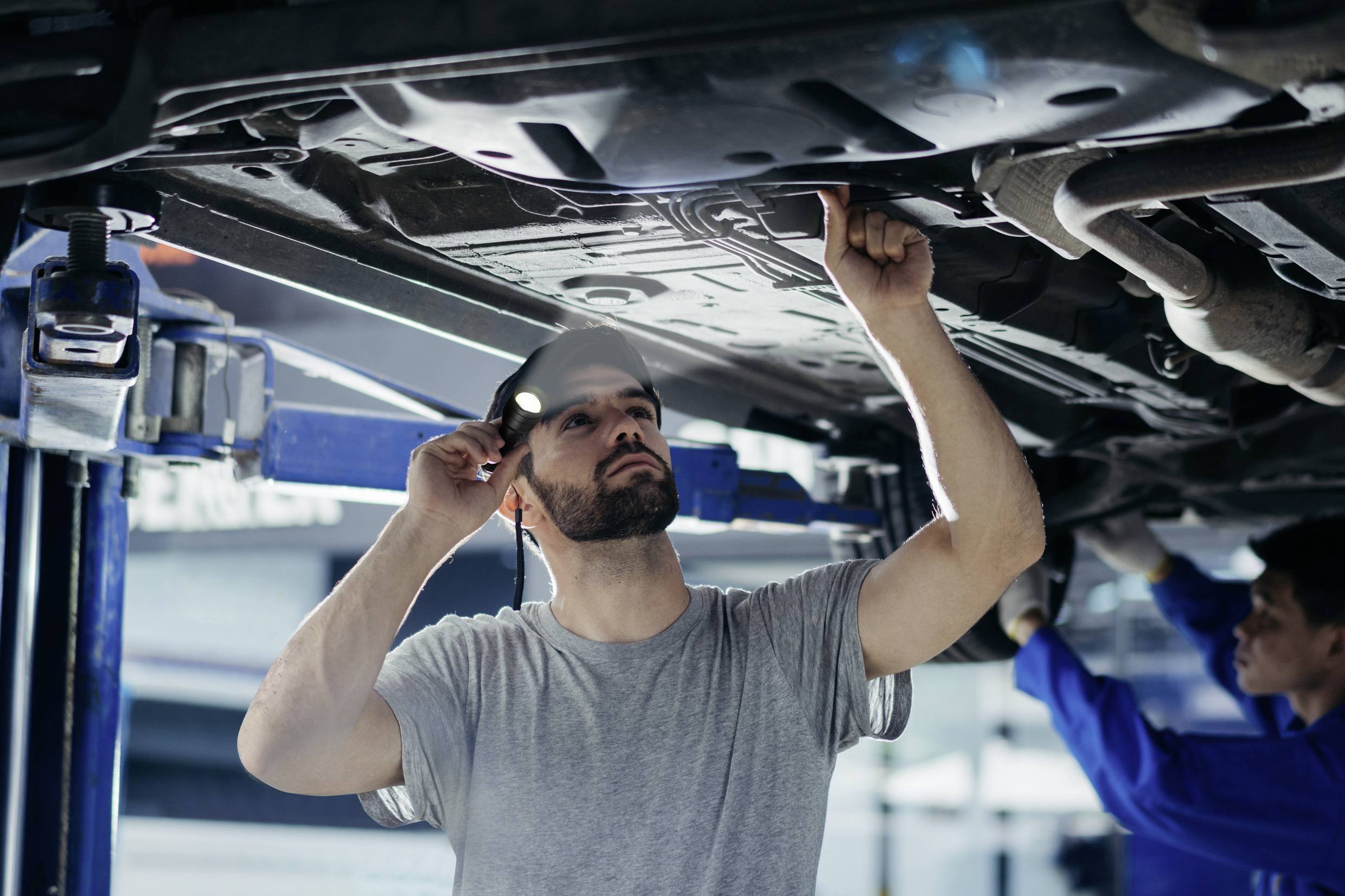 Ein Mechaniker inspiziert den Unterboden eines angehobenen Autos in einer Werkstatt, hält eine Taschenlampe und zeigt auf ein Bauteil.