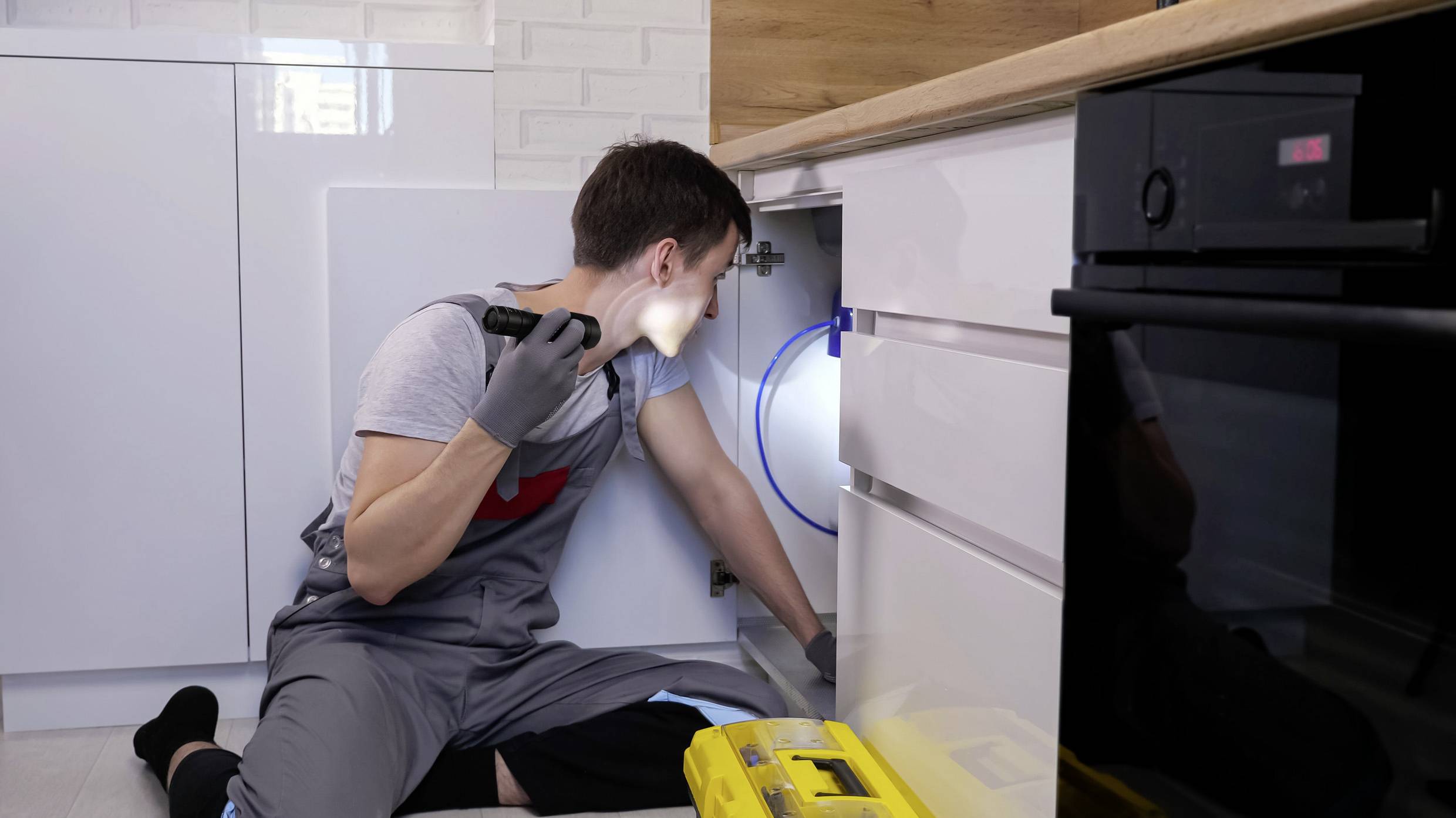 Ein Handwerker in Arbeitskleidung repariert einen Schrank unter der Küche. Er benutzt eine Taschenlampe zur besseren Sichtbarkeit.