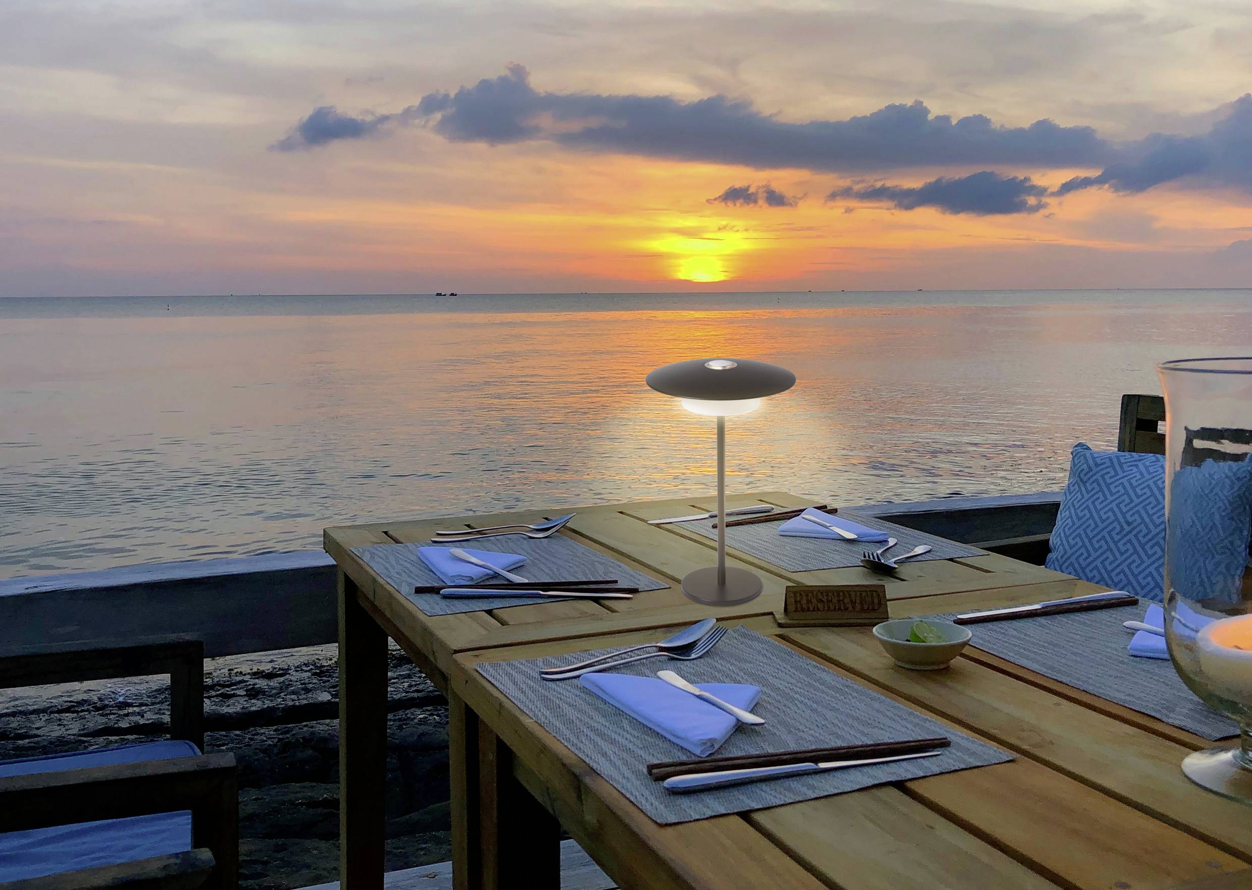 Esstisch für zwei Personen gedeckt mit Besteck und Servietten, mit Blick auf einen friedvollen Sonnenuntergang am Meer, wobei ein orangefarbener Schein auf dem Wasser reflektiert.