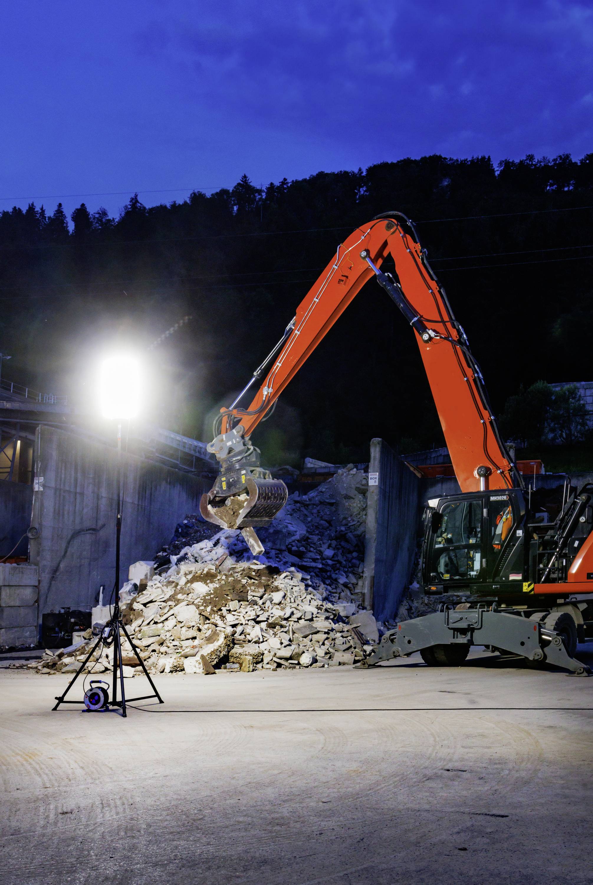 Bagger hebt Steinschutt bei Nacht auf einer Baustelle, beleuchtet von einer hellen Lampe. Im Hintergrund sind Bäume und ein blauer Himmel zu sehen.