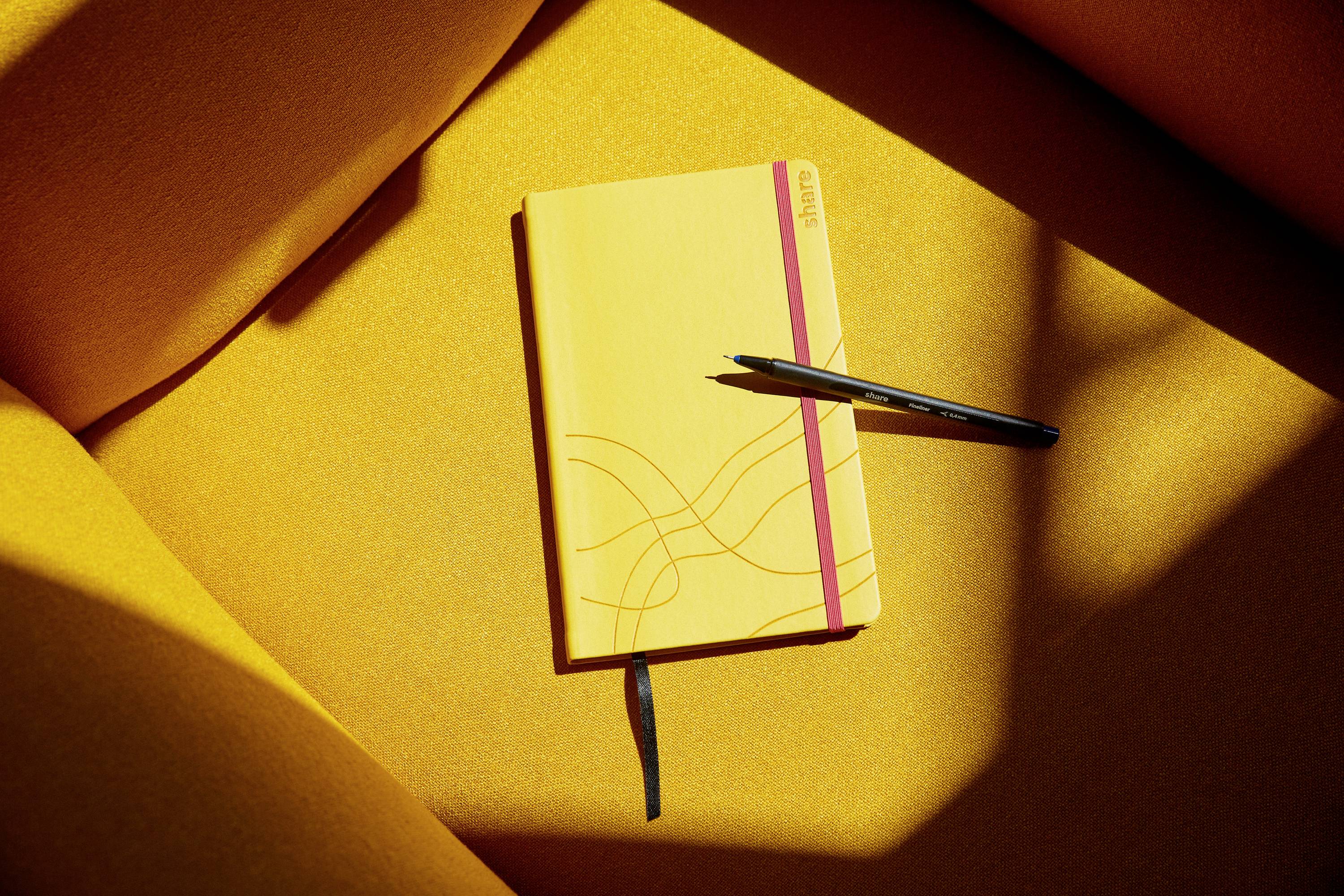 Notizbuch mit orangenem Einband und Stift auf einem leuchtend gelben Sofa, beleuchtet von Sonnenstrahlen.