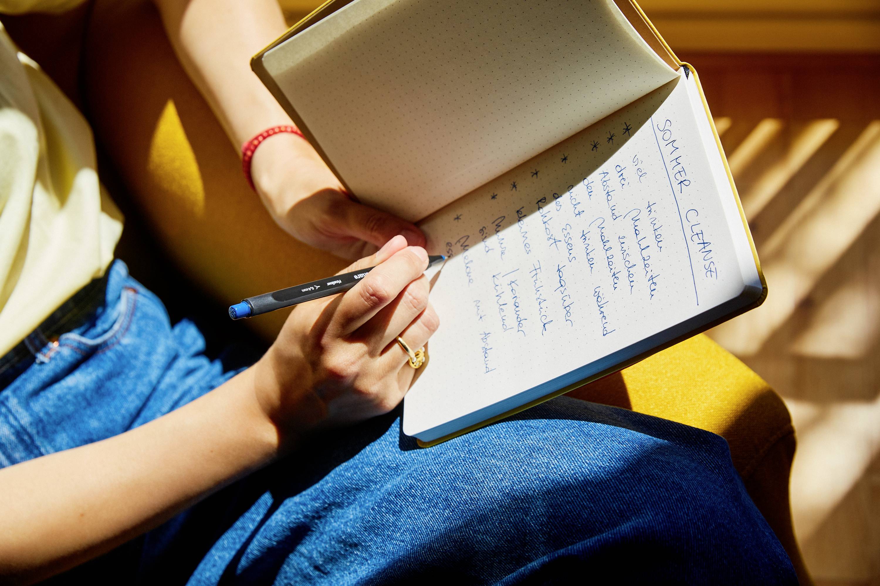 Eine Person sitzt auf einem gelben Stuhl und notiert Sommerpläne in ein aufgeschlagenes Notizbuch. Sonnenlicht fällt auf das Buch.