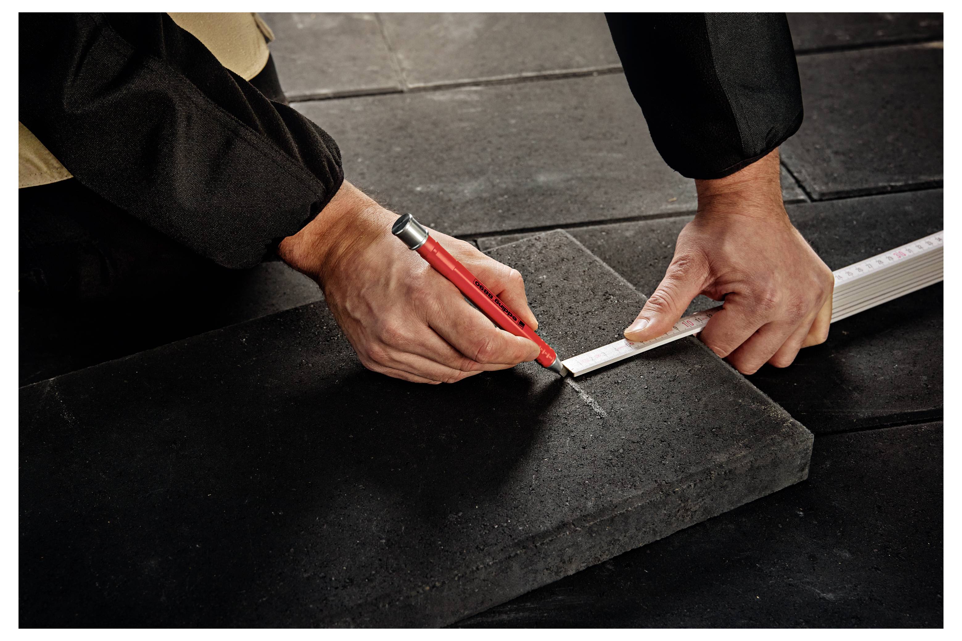 Person, die mit einem Lineal und einem roten Bleistift ein dunkles Dachziegel oder Dachmaterial während der Installation oder Reparatur misst und markiert.