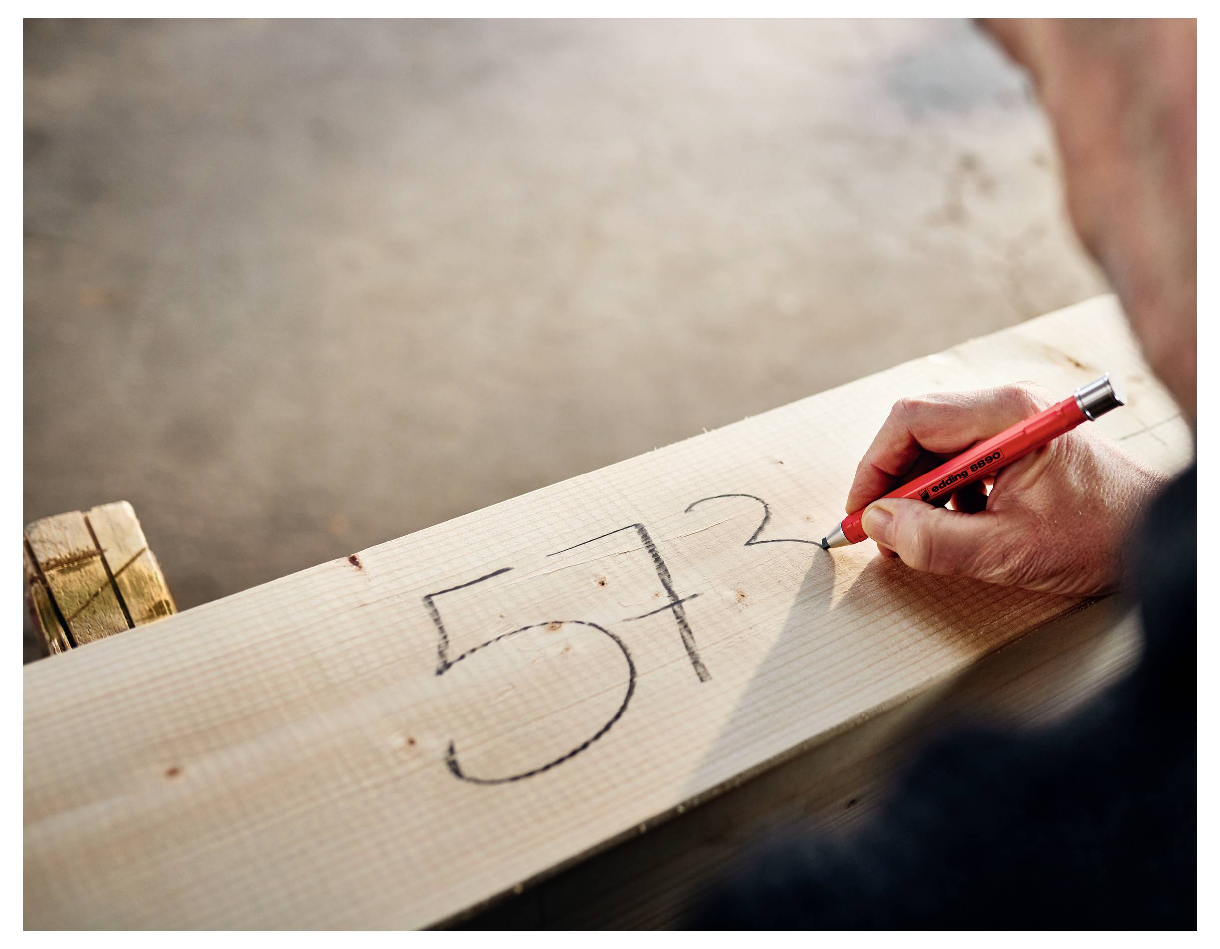 Eine Hand schreibt die Zahl 573 mit einem roten Stift auf ein Stück Millimeterpapier, das auf einer Holzoberfläche liegt.