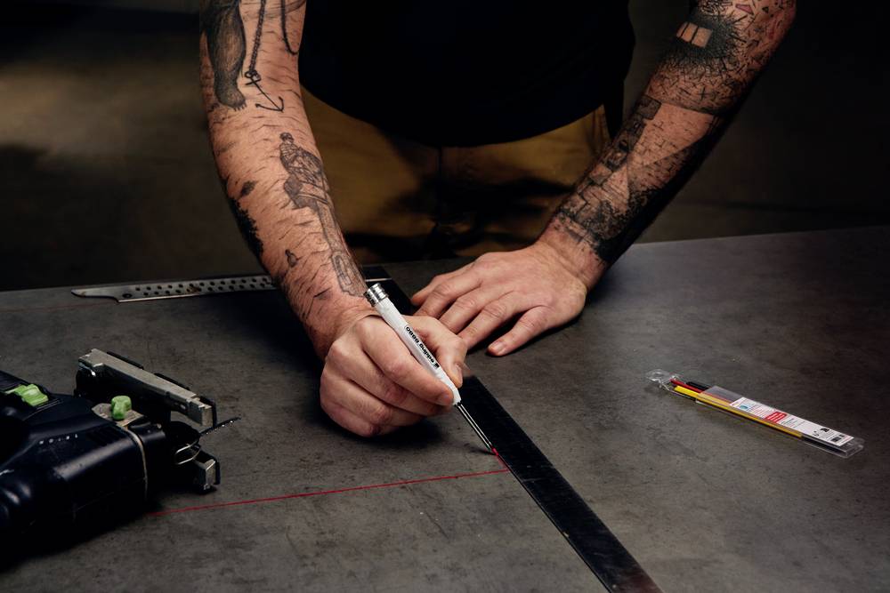 Eine Person mit tätowierten Armen misst auf einem dunkelgrauen Tisch mit einem Lineal und einem Stift. Werkzeuge liegen daneben.