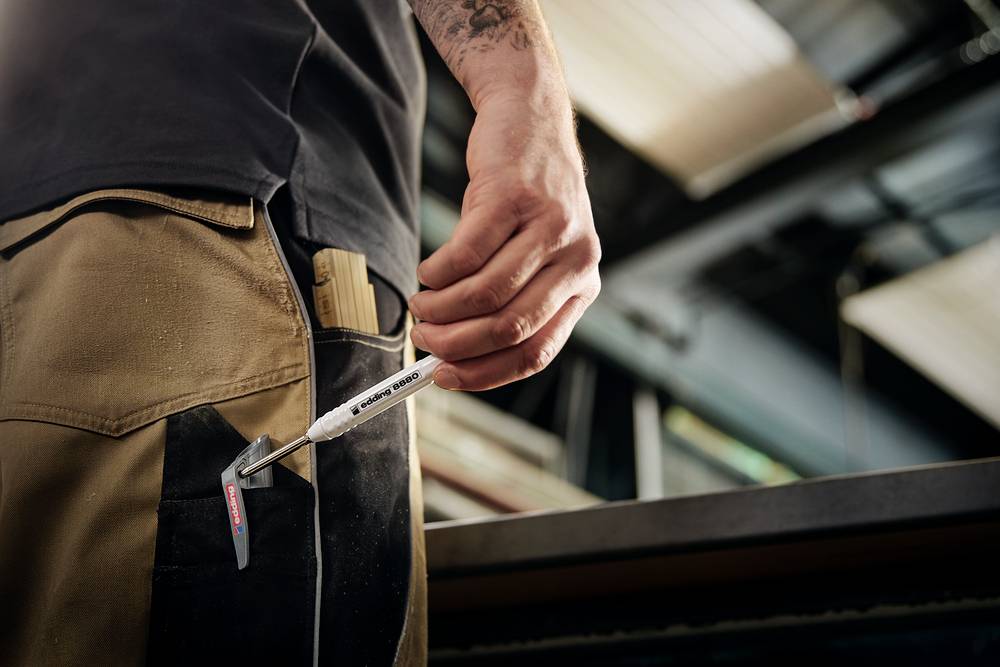 Eine Person in Arbeitskleidung steht in einer Werkstatt. Sie hält einen weißen Stift mit der Aufschrift 'Mess schnell, schreib schnell' in der Hand.
