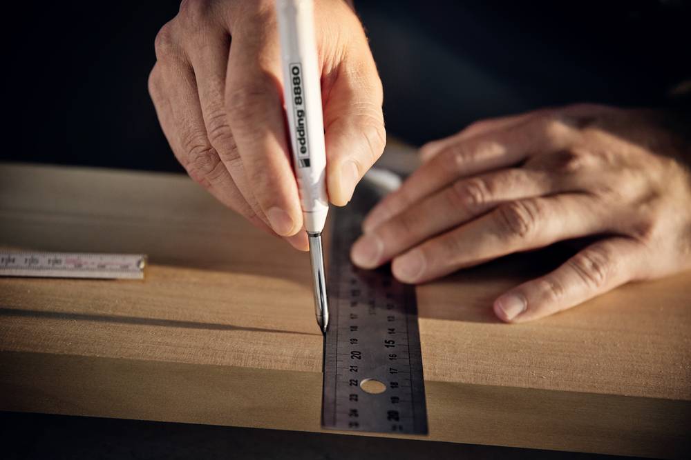 Eine Hand zeichnet mit einem Stift eine Linie entlang eines Metalllineals auf ein Holzbrett. Ein Maßband liegt daneben.