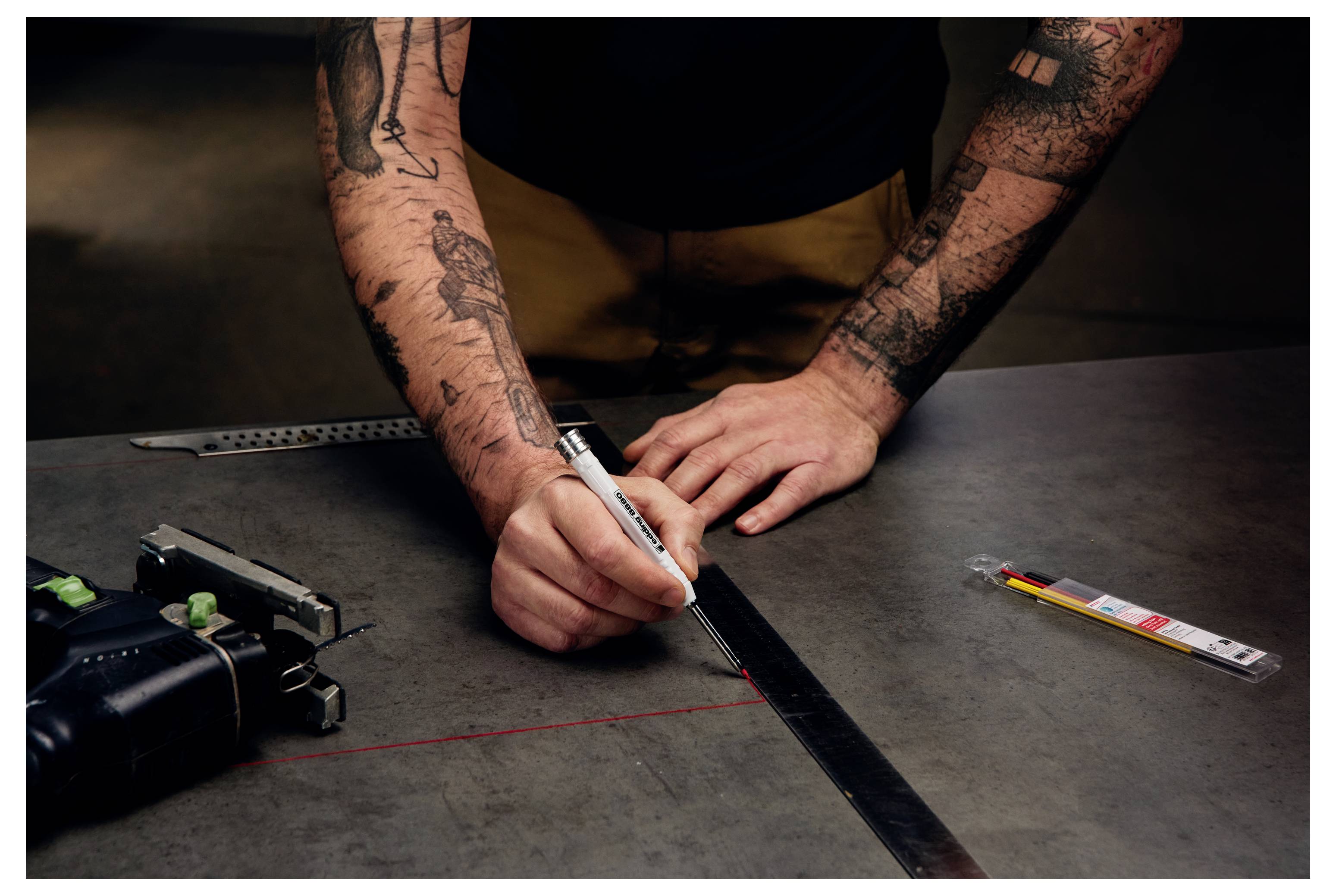 Tätowierte Person, die mit einem Marker eine gerade Linie auf einer Metalloberfläche zeichnet, dabei ein Lineal und ein Messwerkzeug auf einer Werkbank verwendet.