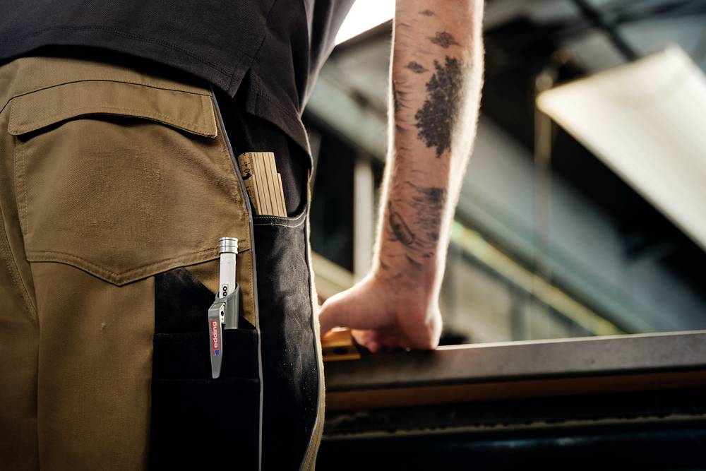 Ein Mensch in Arbeitskleidung lehnt an einer Werkbank. In der Hosentasche stecken Werkzeuge wie ein Bleistift und ein Maßstab.