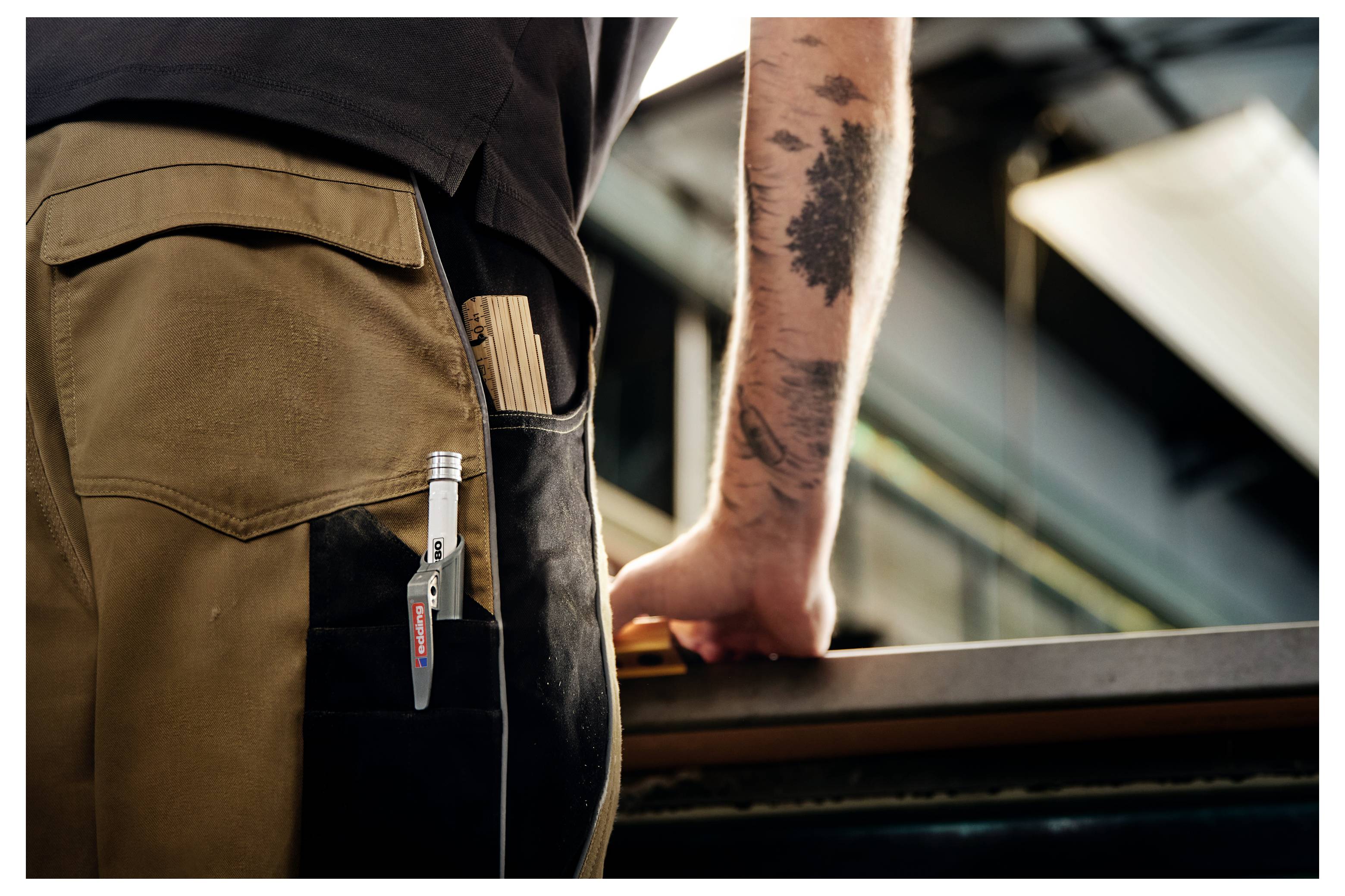 Nahaufnahme eines Arbeiters in beiger Hose mit einem Stift in der Tasche, der einen tätowierten Arm auf einem Geländer in einer Industrieumgebung ablegt.