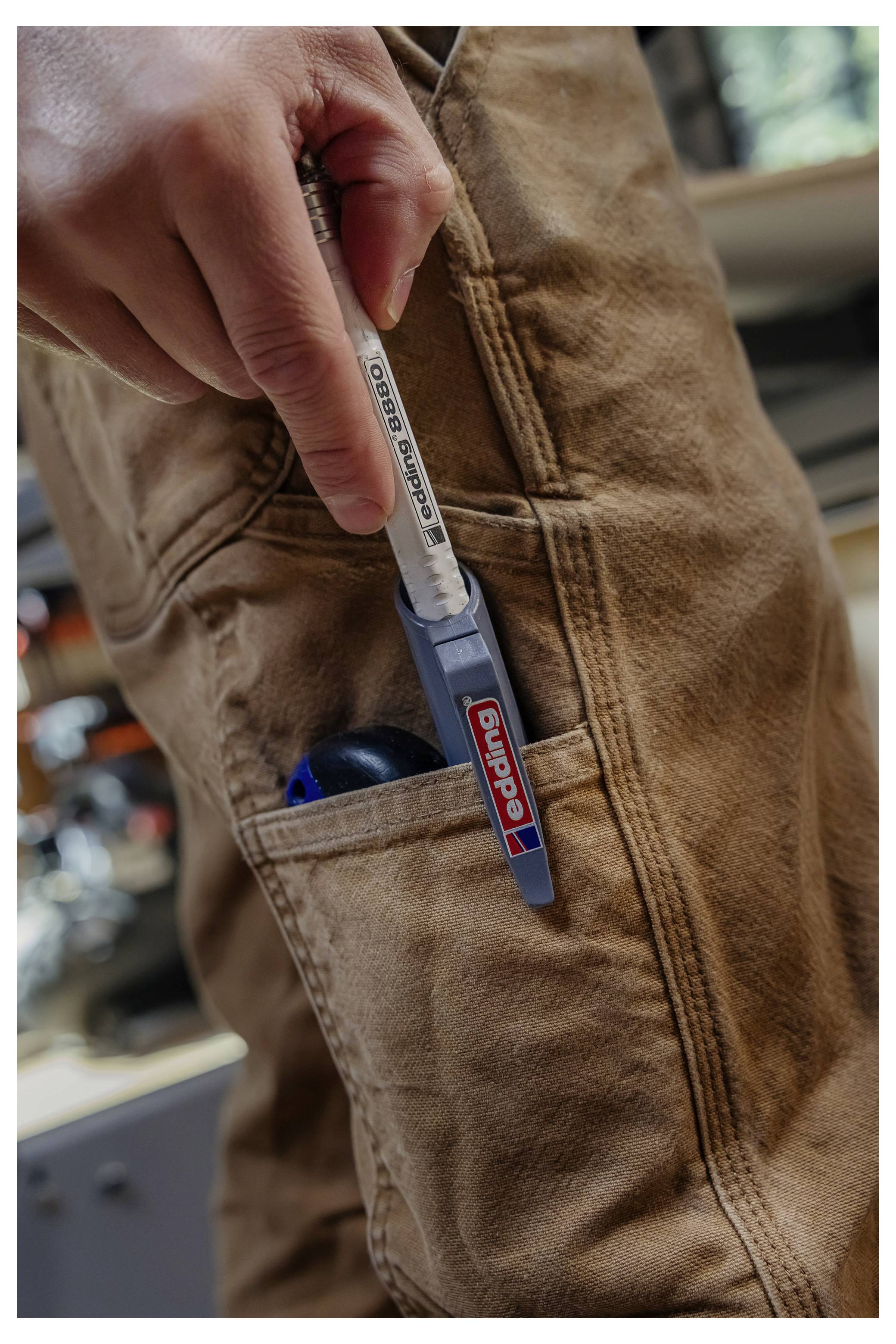 Hand, die einen stiftähnlichen Werkzeug in einer Hosentasche aus brauner Arbeitskleidung hält, wobei ein kleines blaues Utensil außen an der Tasche befestigt ist.