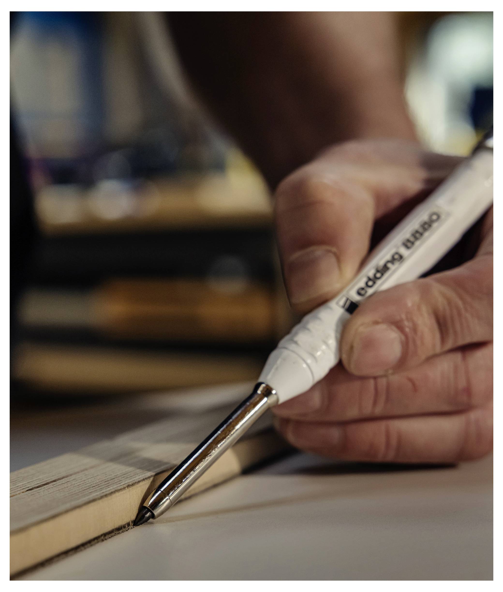 Nahaufnahme einer Hand, die einen weißen Stift verwendet, um eine feine Linie entlang der Kante eines Holzstücks auf einer Arbeitsfläche zu zeichnen.