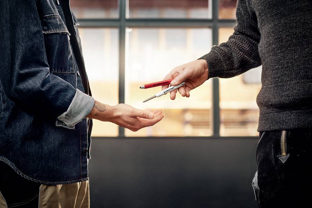 Person übergibt einer anderen Person einen Stift; die Szene findet vor einem Fenster statt.