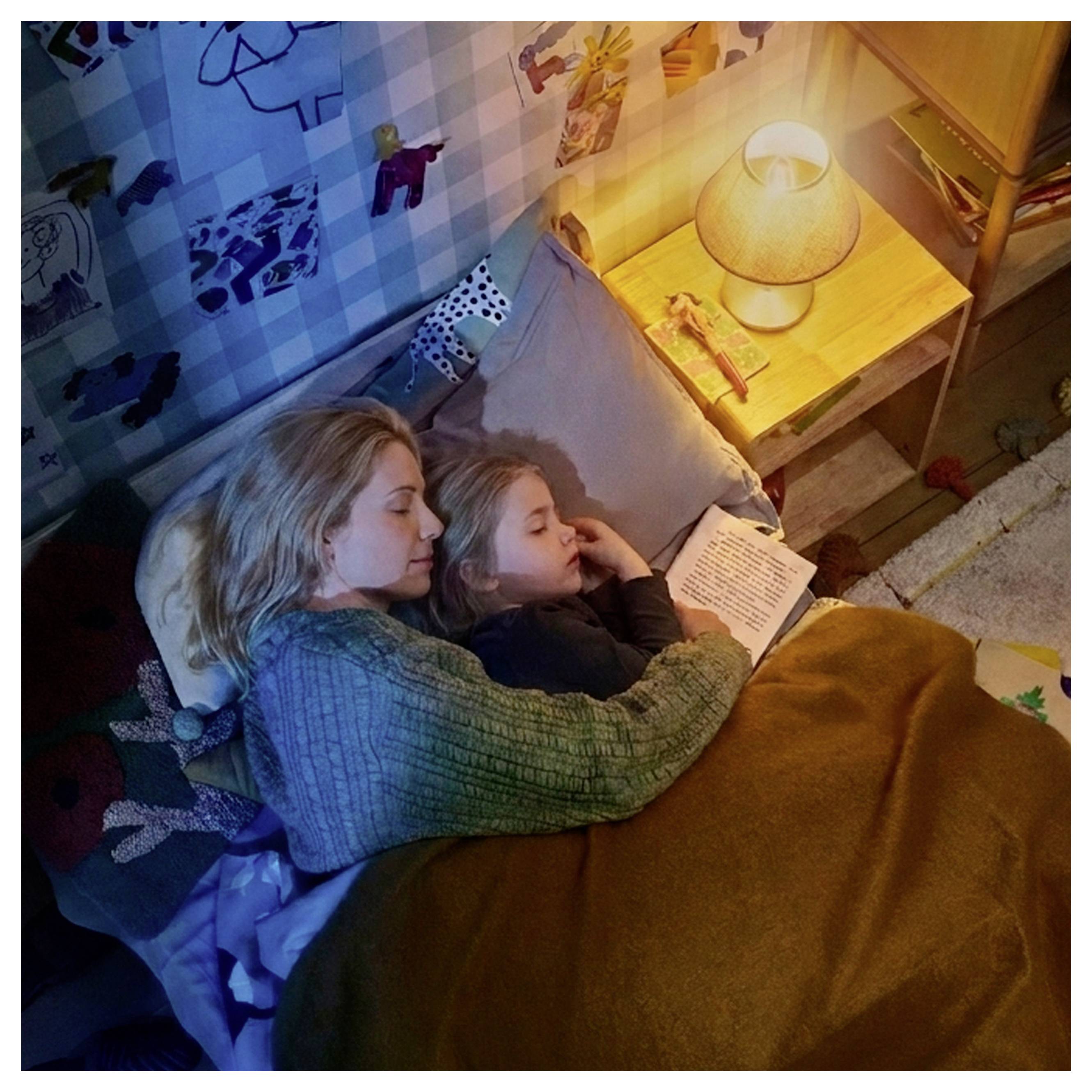 Eine Mutter und ein Kind liegen sich kuscheld im Bett, lesen ein Buch bei einer Nachttischlampe in einem gemütlichen Schlafzimmer.