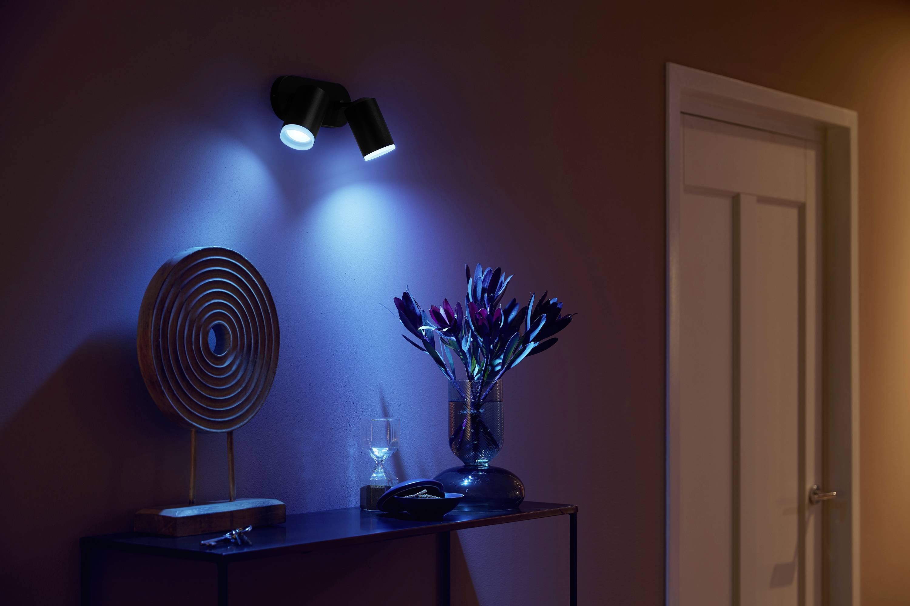 Ein stilvoll beleuchteter Raum mit einer Skulptur und Blumen auf einem Tisch; warmes und blaues Licht erzeugt eine beruhigende Atmosphäre.