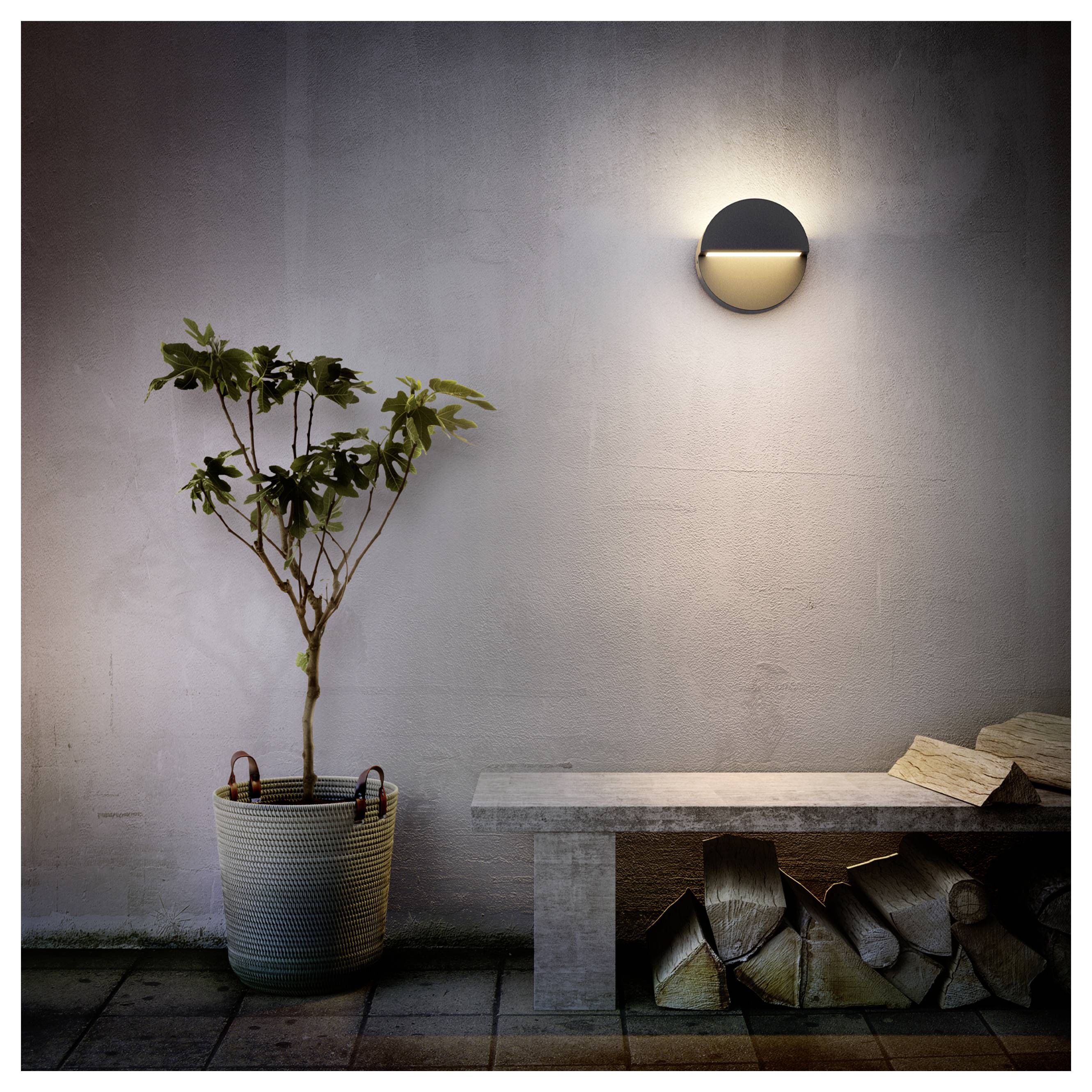 Minimalistischer Terrassenbereich mit einem Baum im Topf, einer Steinbank, gestapeltem Brennholz und einer beleuchteten Wandleuchte an einer einfachen Wand.