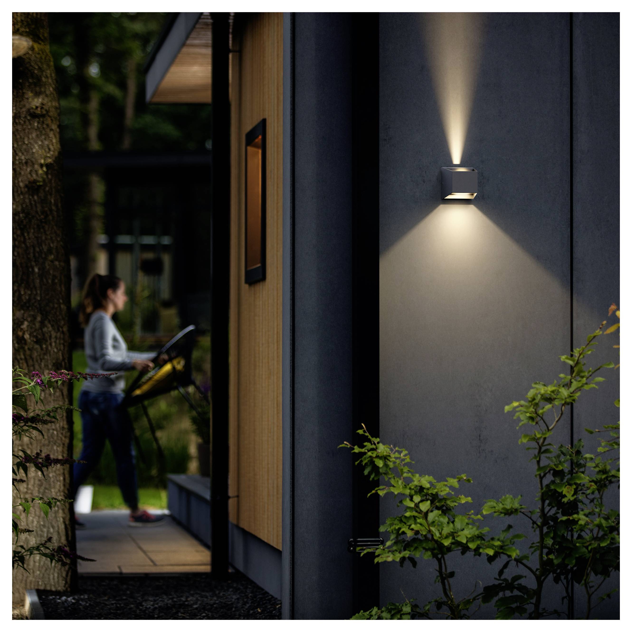 Nächtliche Außenaufnahme eines modernen Hauses, beleuchtet durch eine Wandleuchte, mit einer Frau, die im Hintergrund auf der Veranda ein Laufband oder Trainingsgerät benutzt.