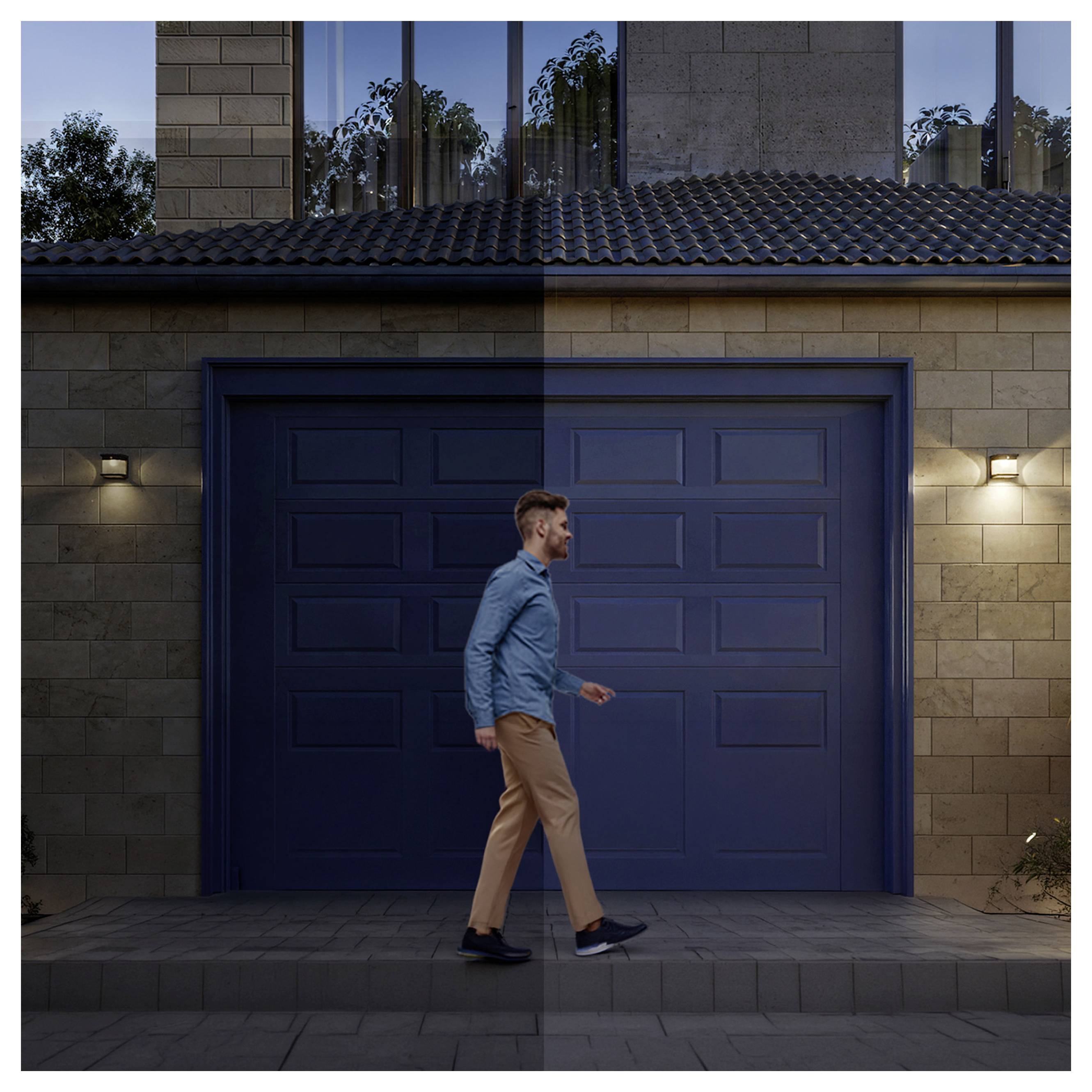 Ein Mann geht an einer modernen Garage mit geschlossenem blauen Tor eines zeitgenössischen Hauses in der Dämmerung vorbei, wobei die Außenwandleuchten eingeschaltet sind.