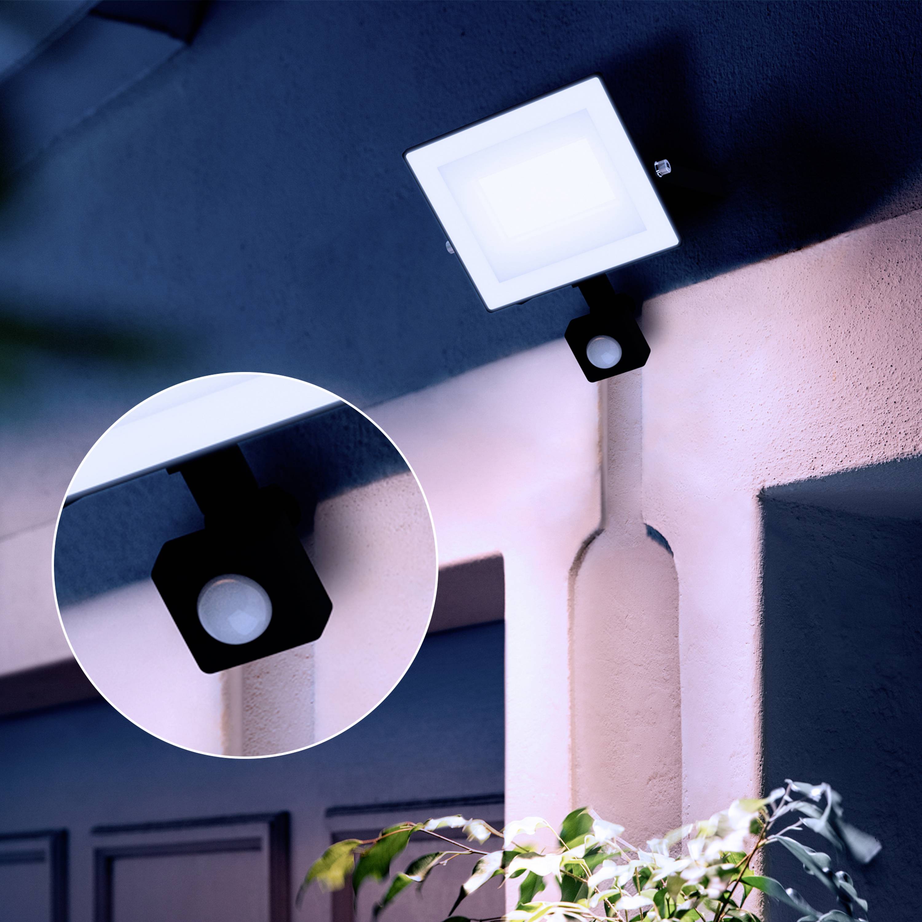 Erleuchtete Außenlampe mit Bewegungsmelder an einer Wand neben einer Tür, in einer nächtlichen Szene.