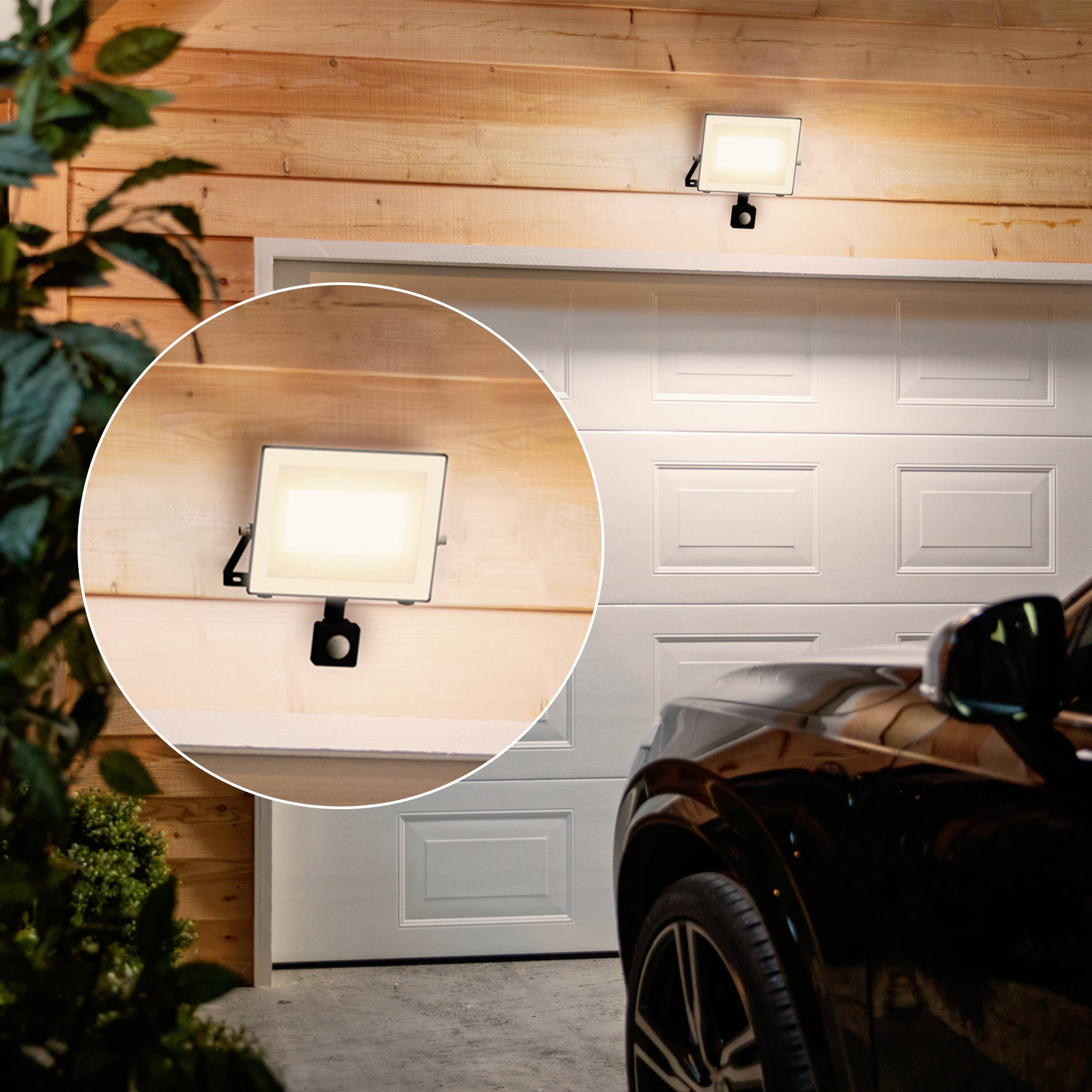 Ein heller Flutlichtstrahler ist über einer Garage montiert, beleuchtet den Raum davor. Im Vordergrund ist ein Auto sichtbar.