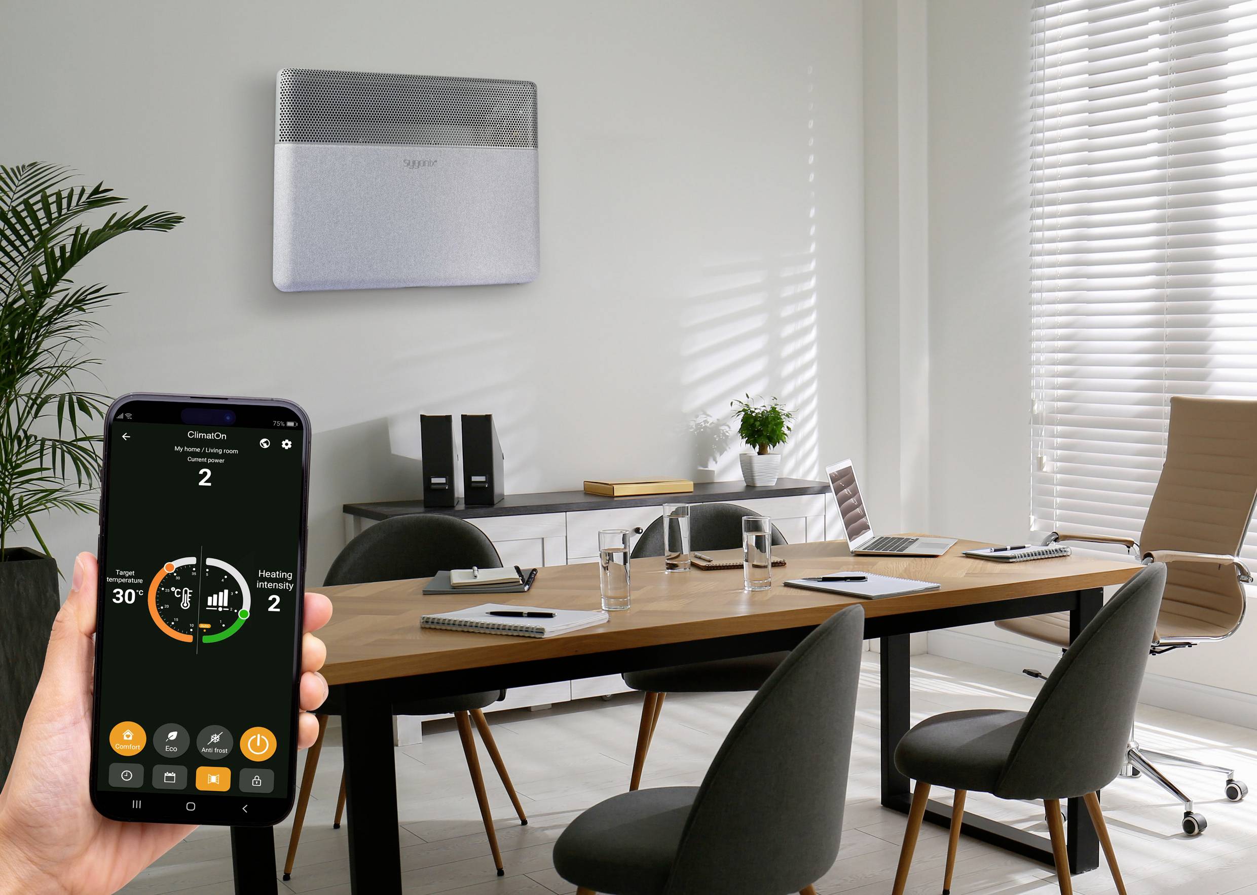 Büro mit Tisch und Stühlen, im Hintergrund ein Heizgerät an der Wand. Eine Hand hält ein Smartphone mit geöffneter Heizungs-App.