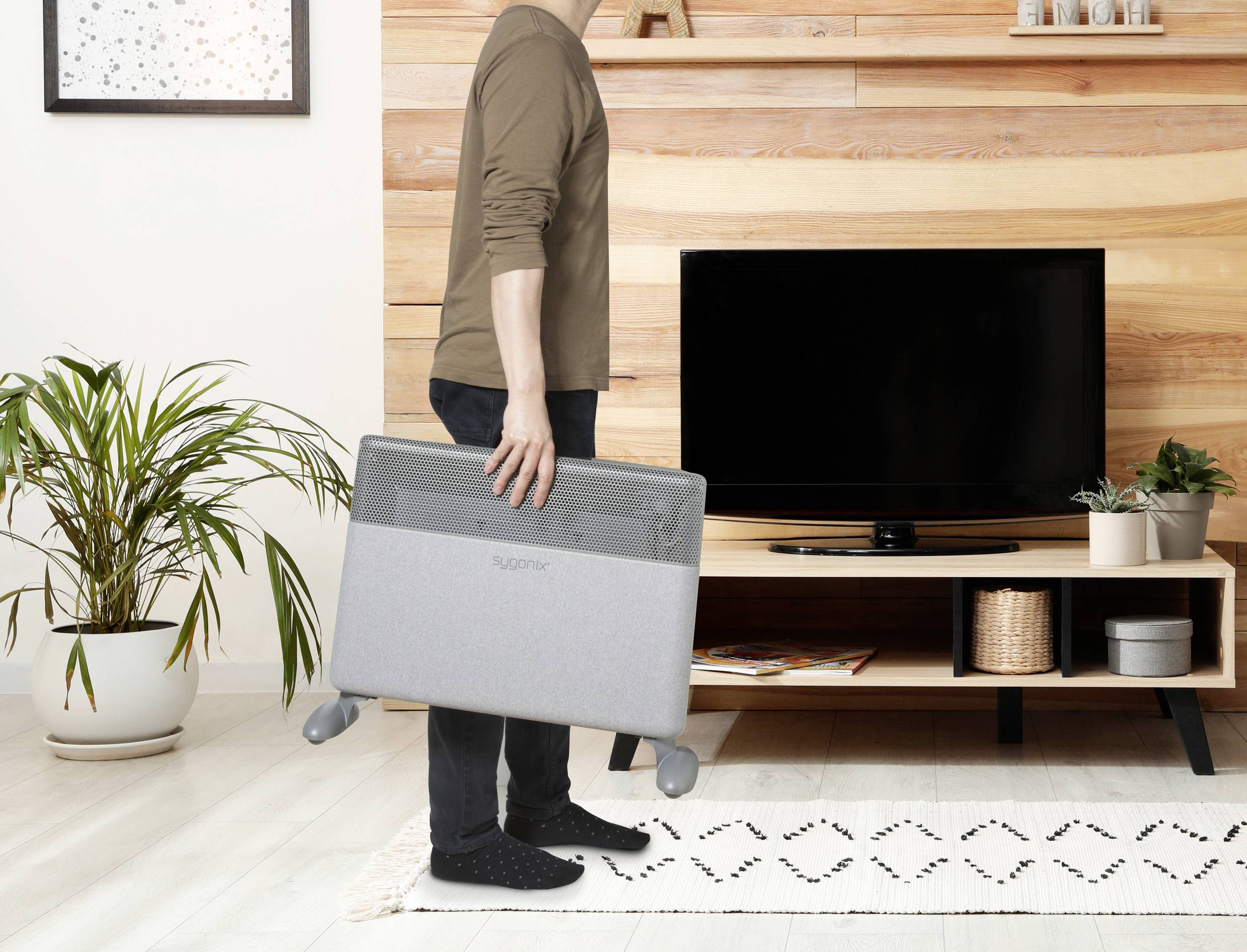 Eine Person trägt einen grauen Heizkörper in einem modernen Wohnzimmer mit Holzverkleidung und einem Fernseher.