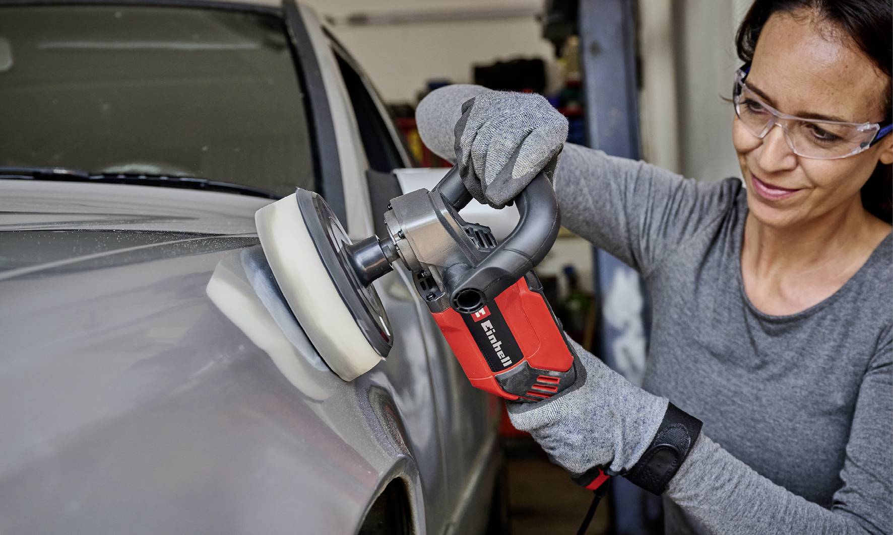 Eine Person poliert mit einer Elektropoliermaschine die Oberseite eines grauen Autos, trägt Schutzbrille und Handschuhe.