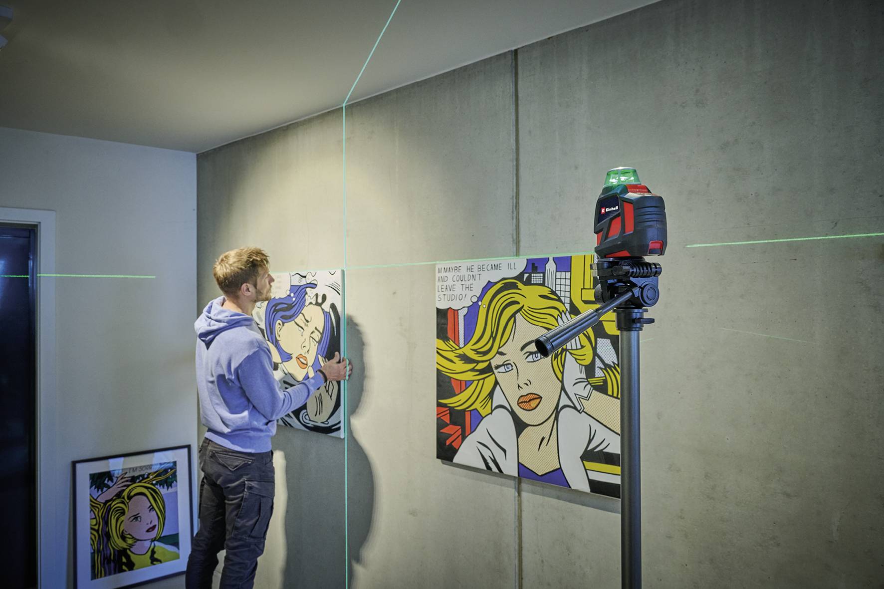 Ein Mann hängt ein Bild an einer Wand auf, unterstützt durch ein grünes Lasergerät zur Ausrichtung. Im Hintergrund sind weitere Gemälde sichtbar.