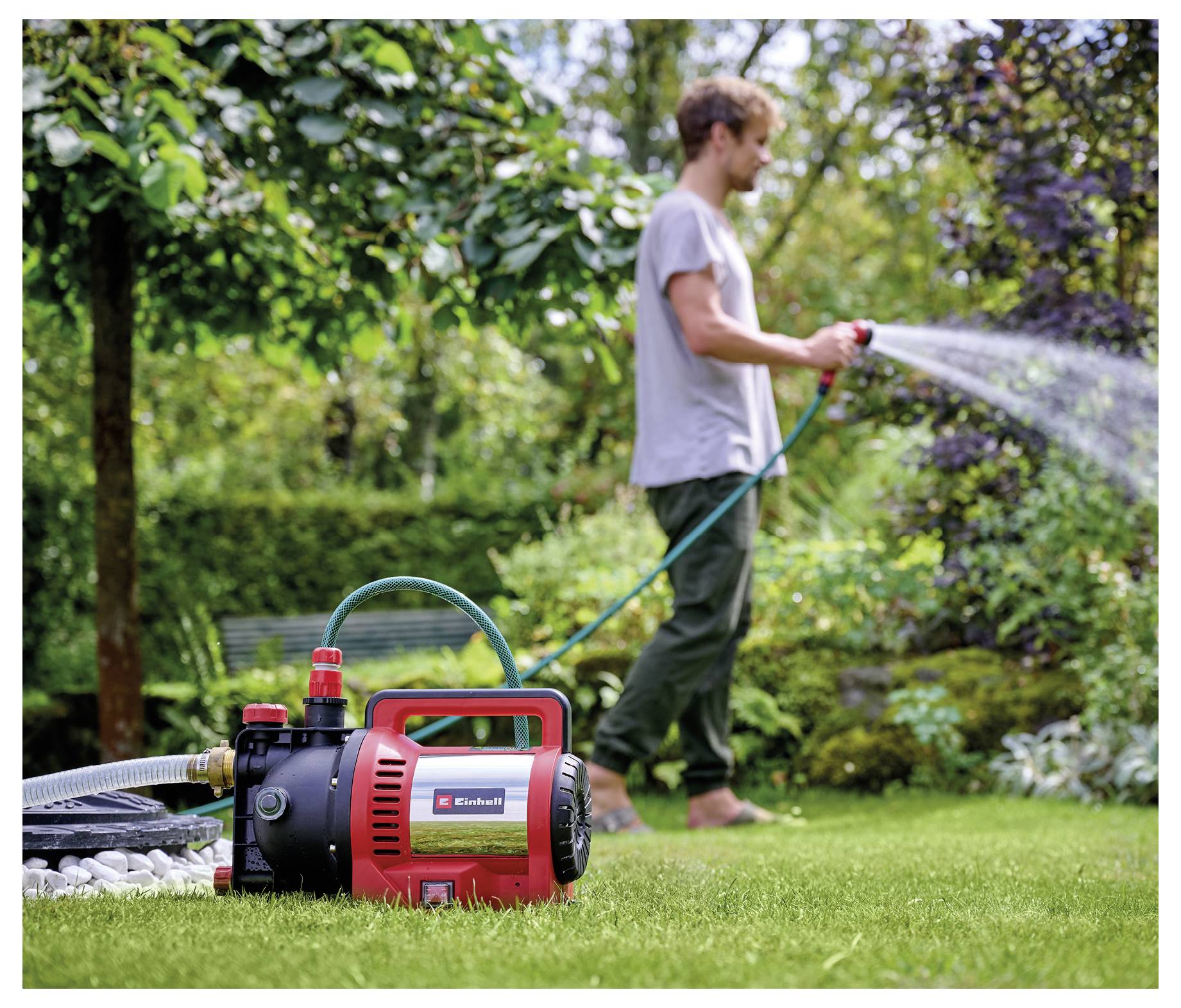 Portable garden pump in the foreground spraying water through a hose as a man waters plants in a backyard garden.