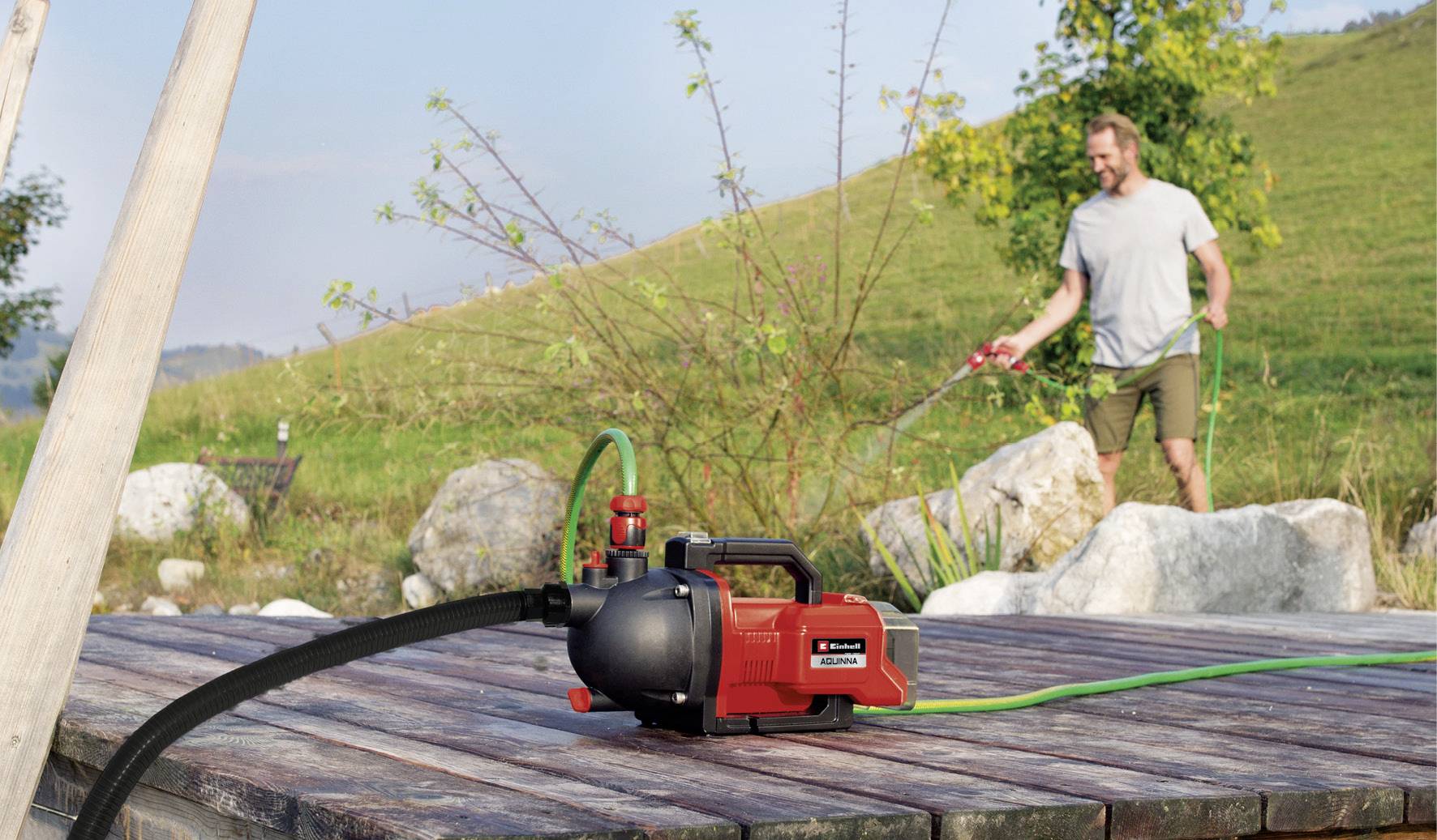 Eine Person bewässert mit einem Gartenschlauch Pflanzen in einem Garten. Eine Wasserpumpe ist prominent im Vordergrund zu sehen.