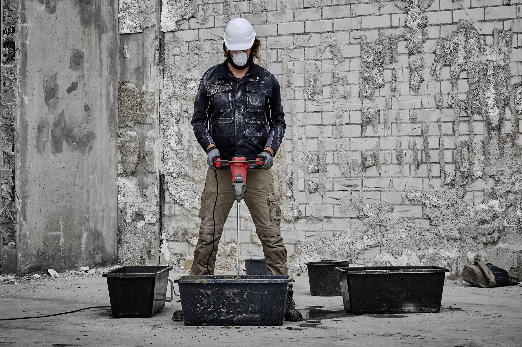 Eine Person mit Schutzhelm und Maske verwendet einen Presslufthammer auf einer Baustelle, umgeben von mehreren großen Behältern.