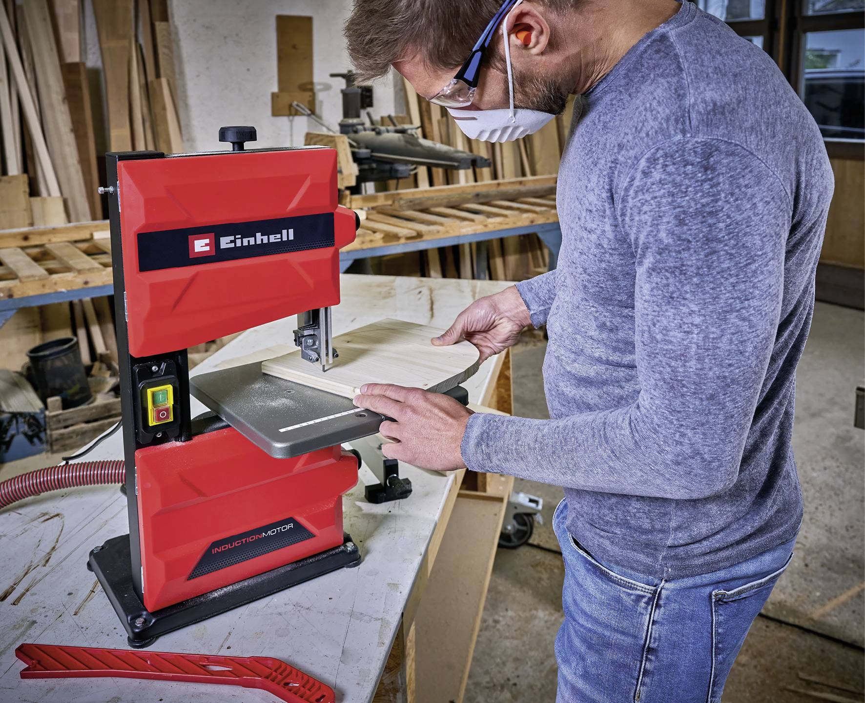 Ein Mann arbeitet mit einer roten Bandsäge in einer Werkstatt. Er trägt Schutzbrille und Maske, schneidet ein Stück Holz.