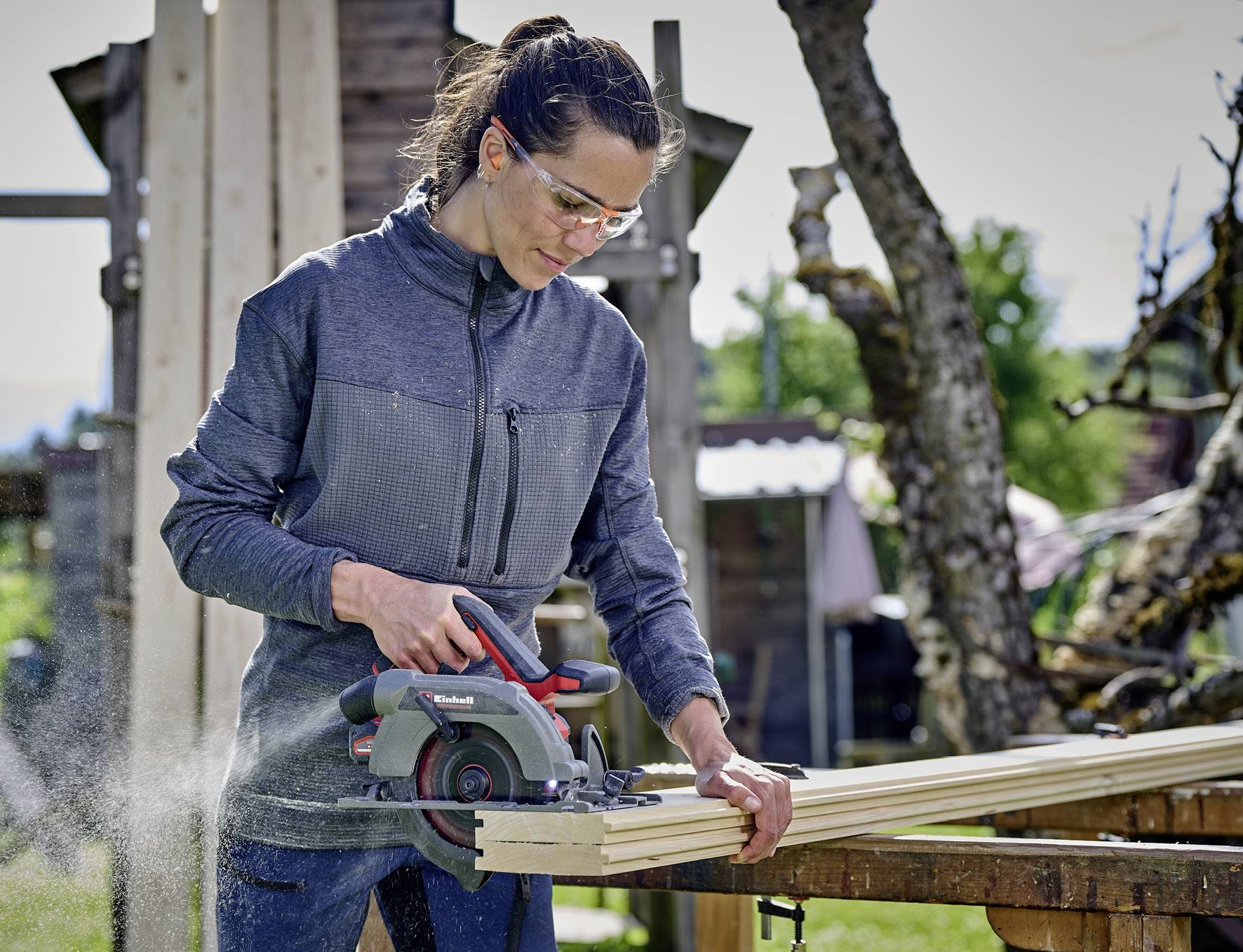 Eine Frau mit Schutzbrille arbeitet draußen mit einer Handkreissäge, um Holzlatten zuzuschneiden.