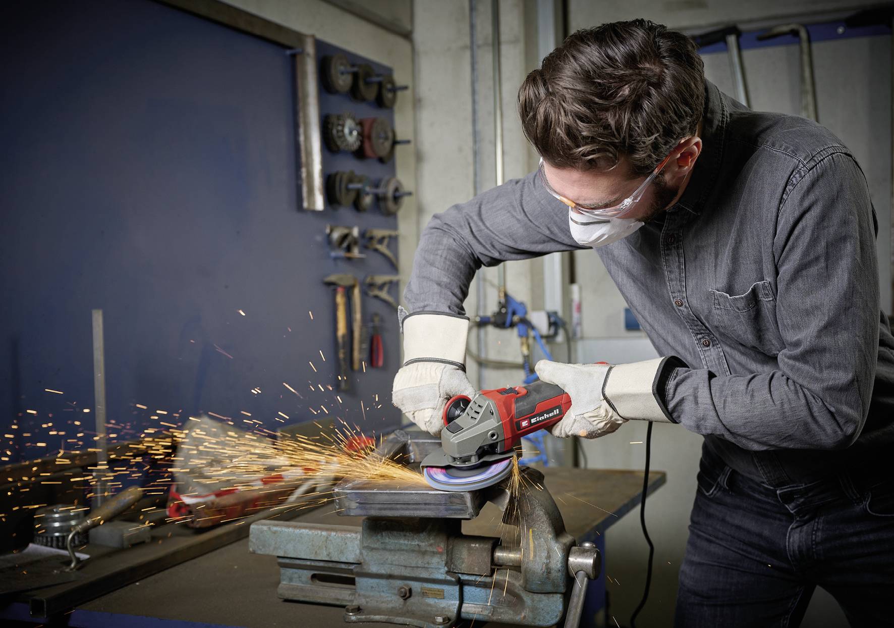 Ein Mann schleift mit einer Schleifmaschine Metall in einer Werkstatt. Er trägt Schutzbrille, Maske und Handschuhe. Funken fliegen.