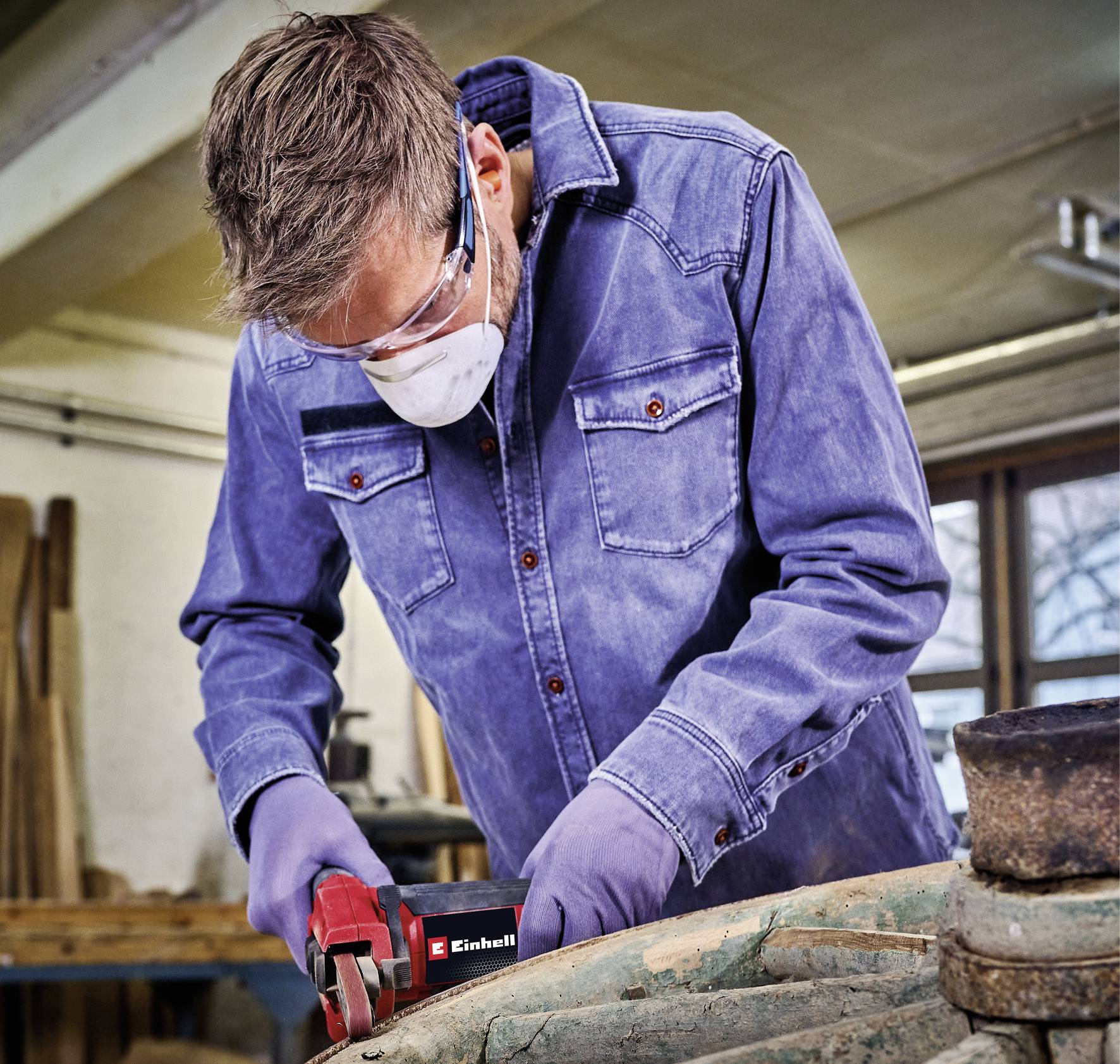 Ein Mann in Schutzbrille und Maske arbeitet in einer Werkstatt mit einem Elektrowerkzeug an einem Holzstück.