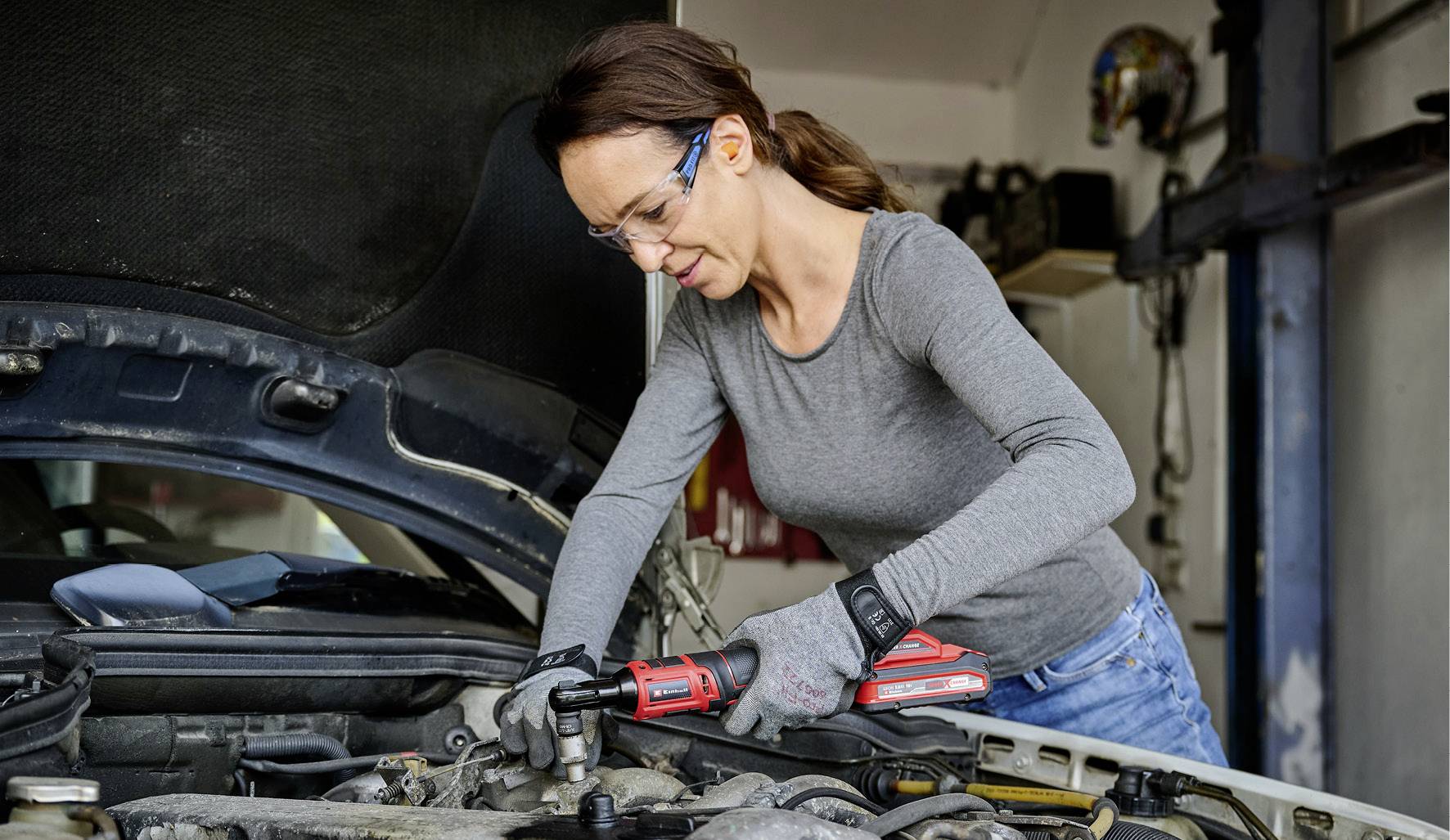 Eine Frau repariert einen Autoreifen in einer Werkstatt. Sie trägt Schutzbrille und Handschuhe und benutzt ein Werkzeug.