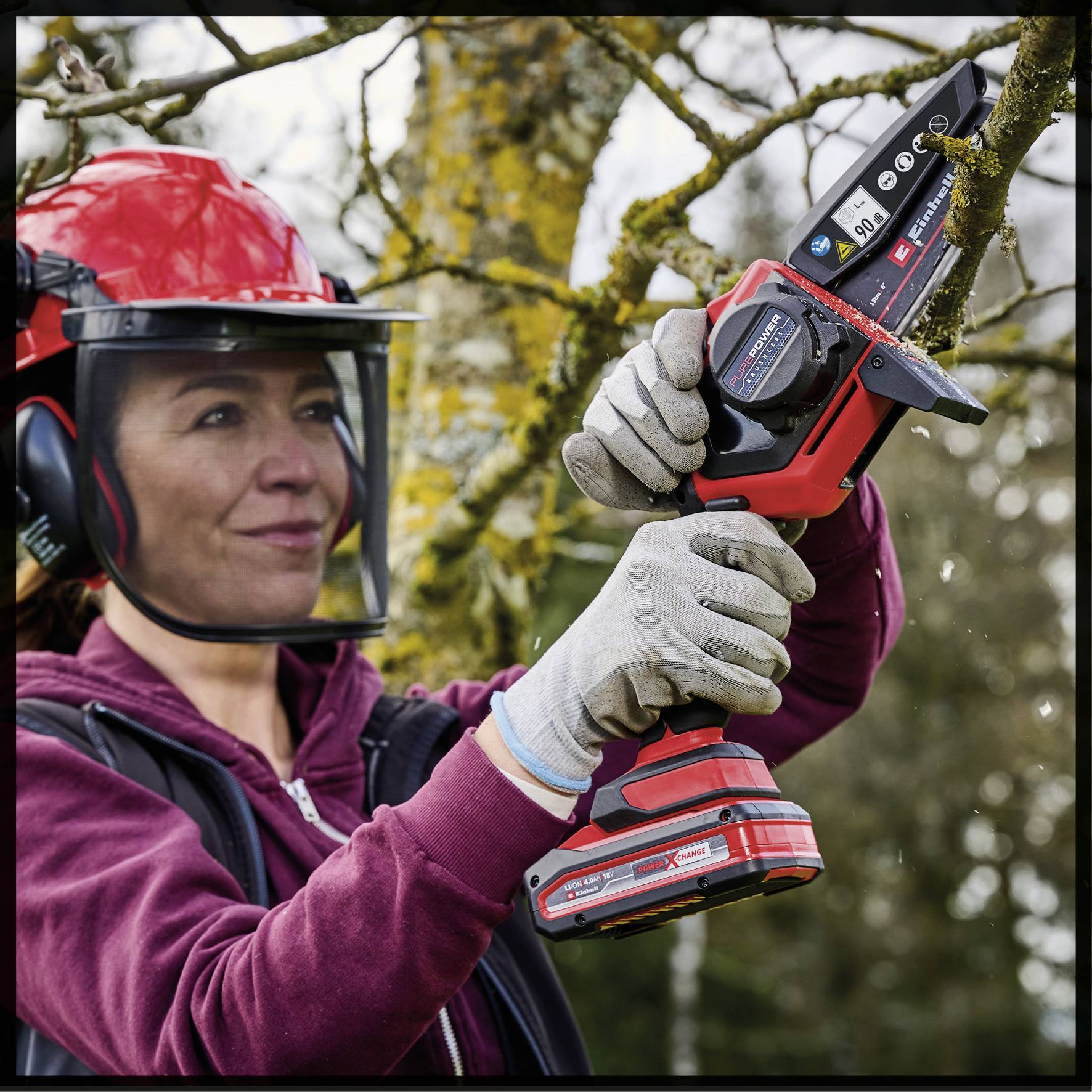 Eine Frau mit Helm und Schutzbrille benutzt eine akkubetriebene Motorsäge, um Äste in einem Baum zu schneiden. Sie trägt rote Arbeitskleidung und Handschuhe.