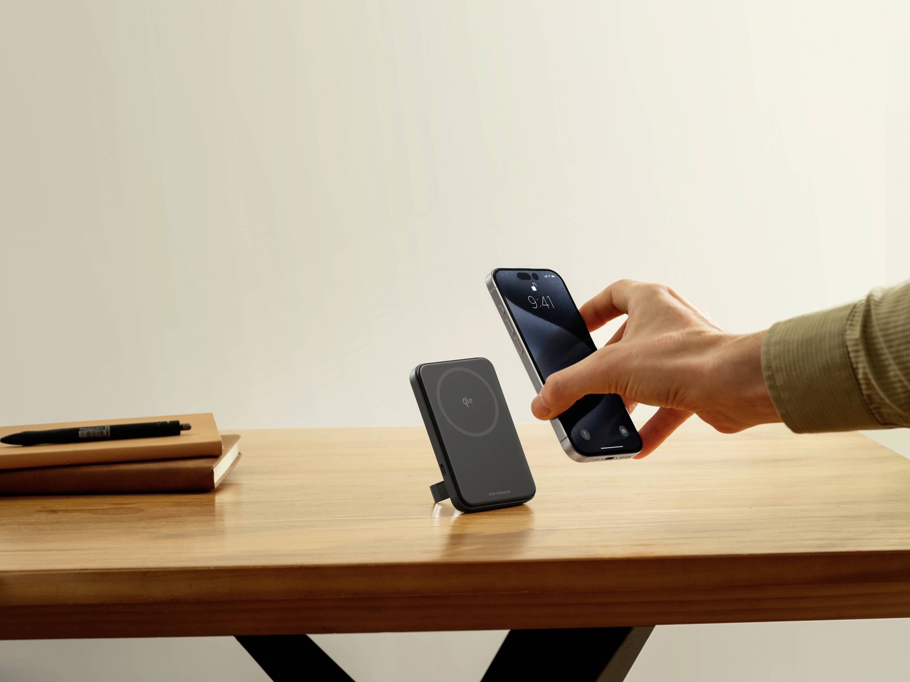 Hand hält Smartphone über kabellose Ladestation auf Holztisch. Im Hintergrund Notizbuch und Stift.