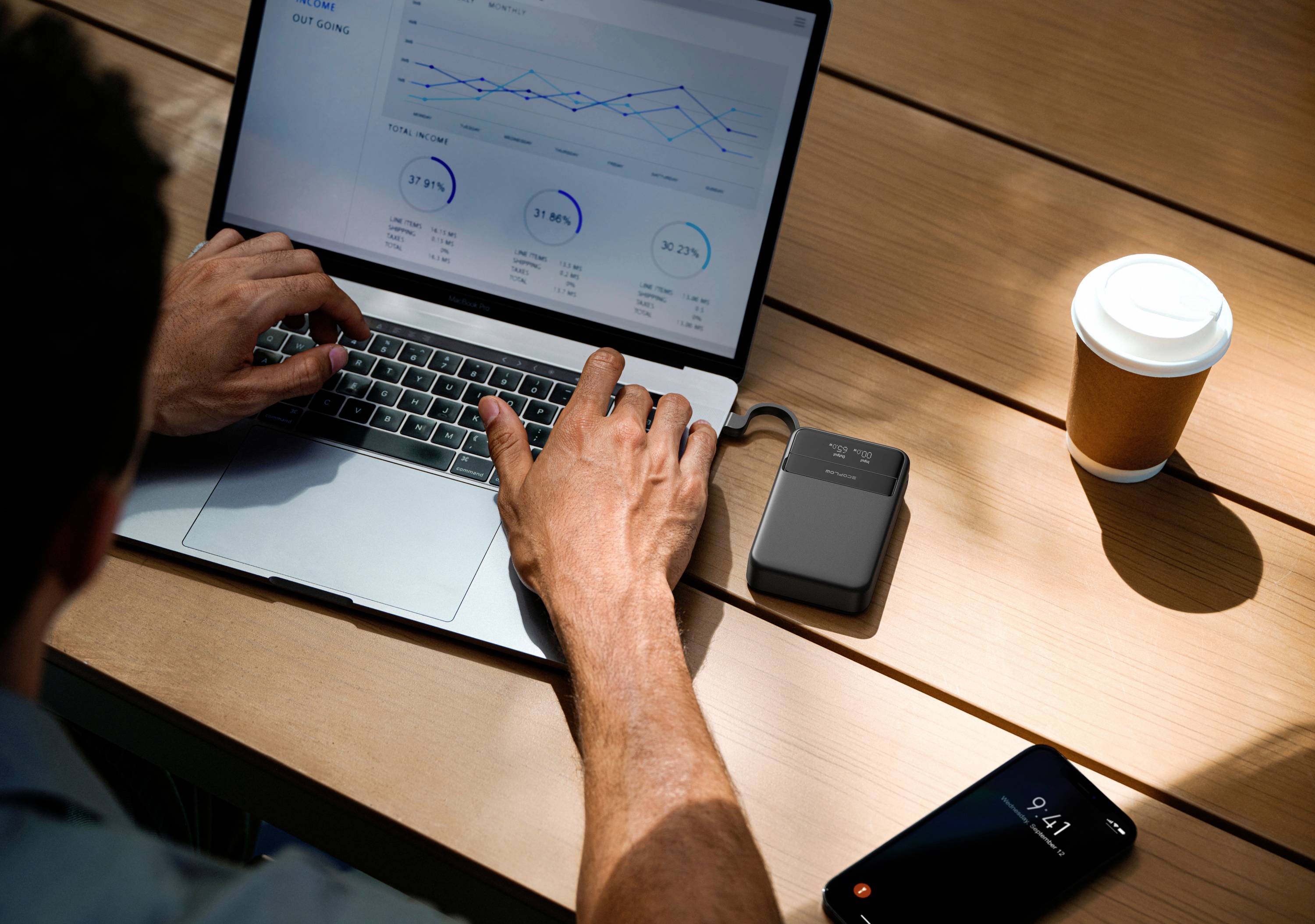 Person arbeitet an einem Laptop mit Diagramm auf dem Bildschirm; daneben Smartphone, Kaffeebecher und externer Akku auf einem Holztisch.
