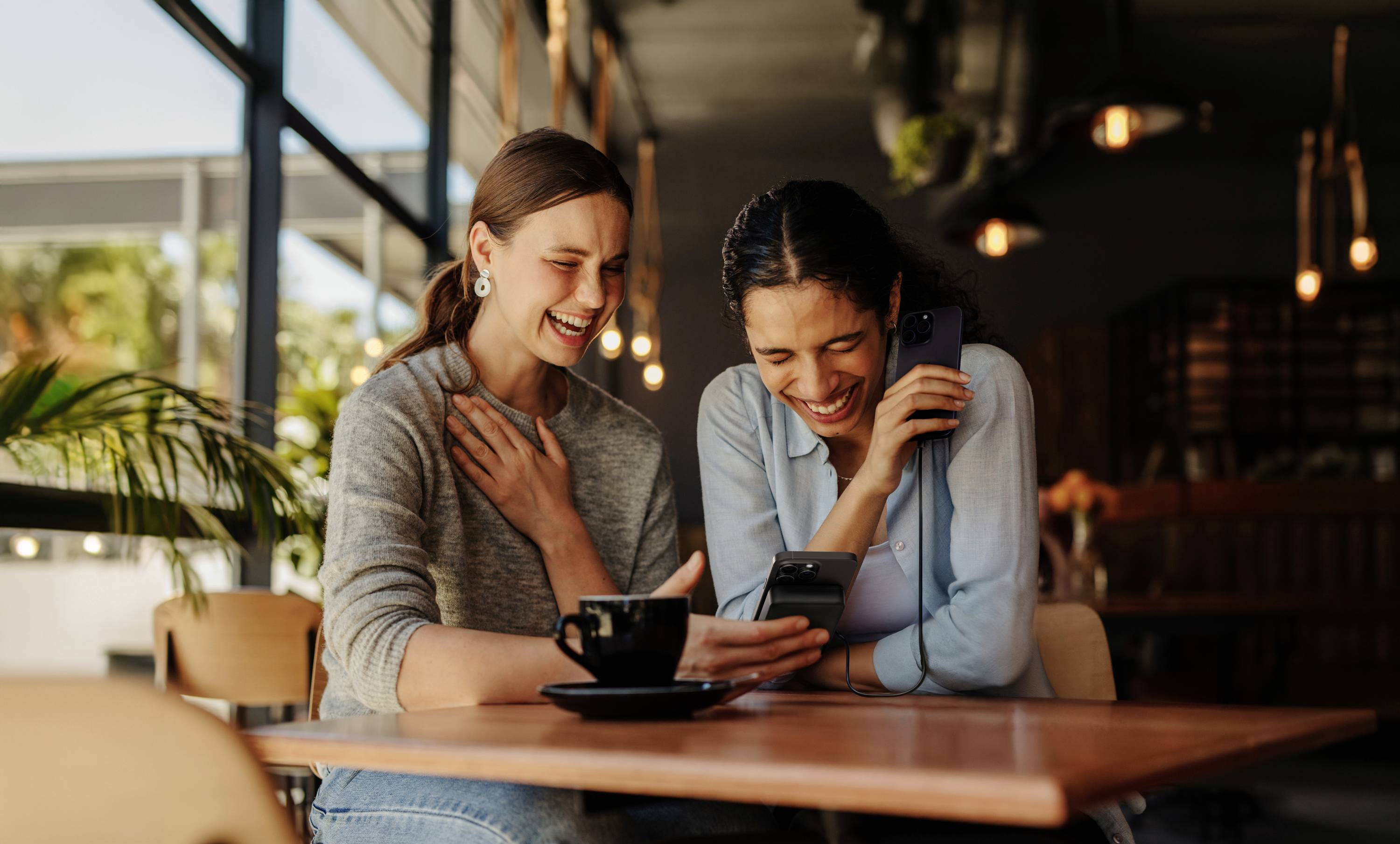 Zwei lachende Frauen sitzen in einem Café, schauen gemeinsam auf ein Smartphone und wirken fröhlich und entspannt.