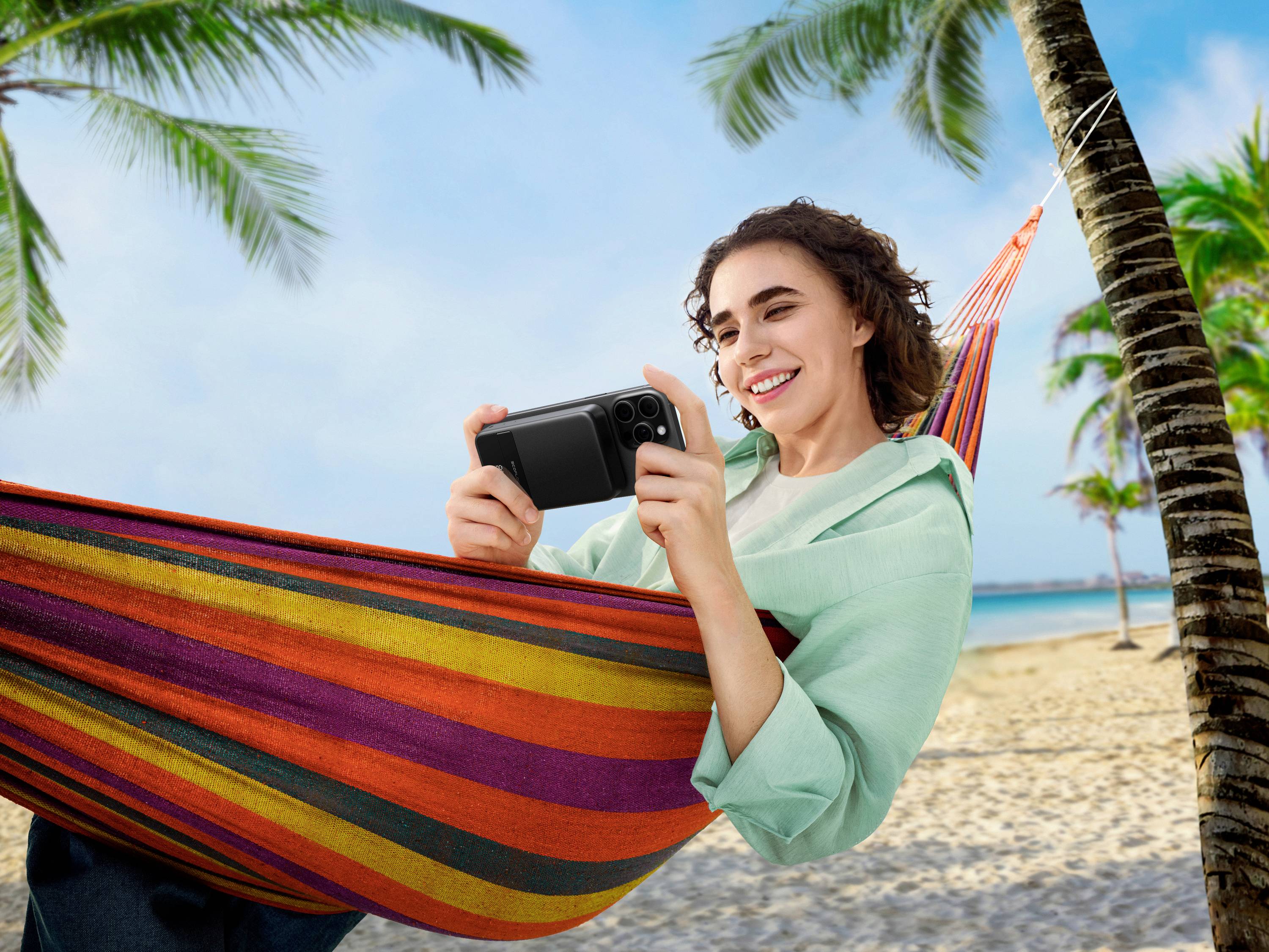 Frau liegt in einer bunten Hängematte am Strand und lächelt, während sie auf ihr Smartphone schaut; Palmen im Hintergrund.