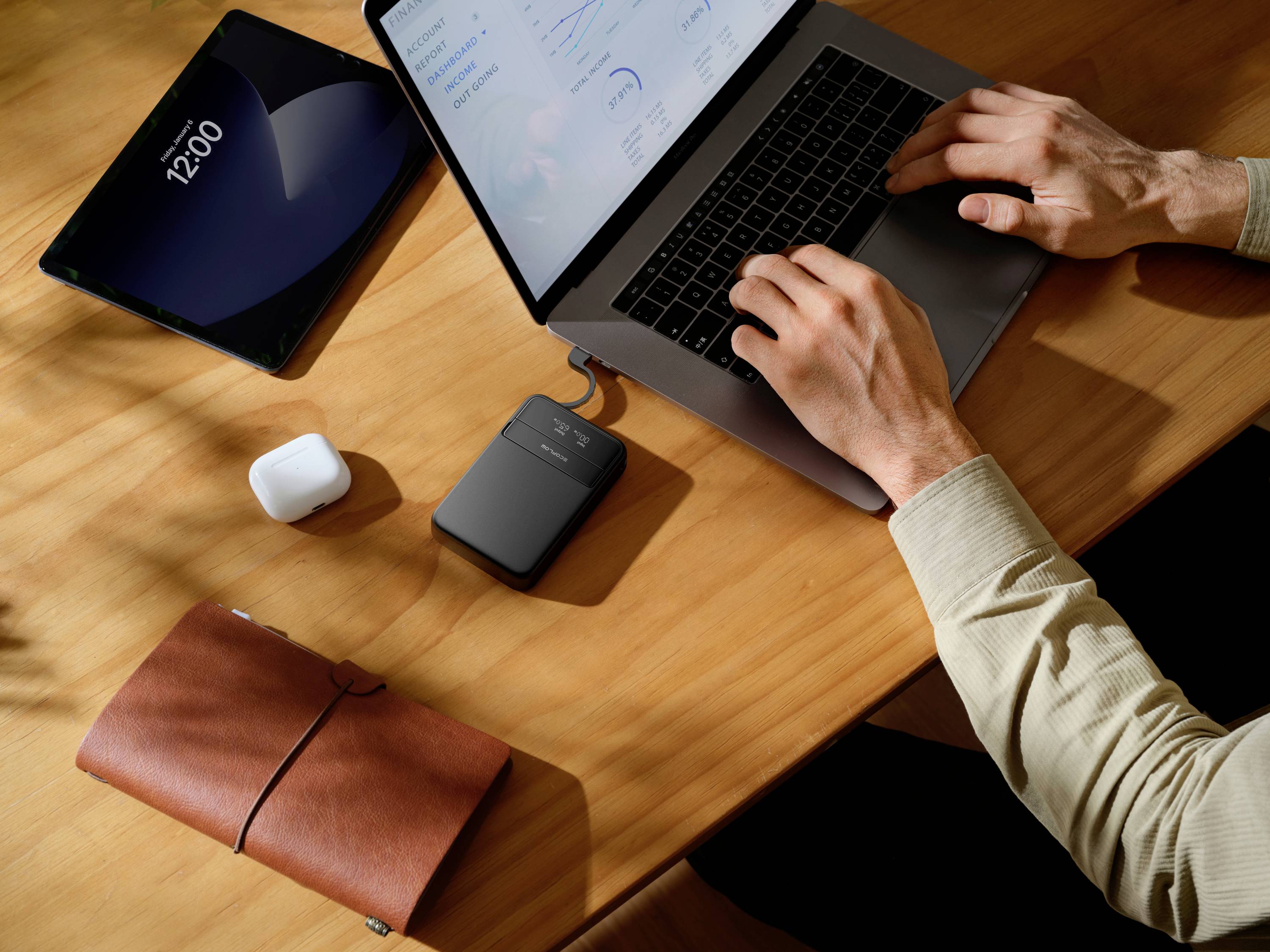 Eine Person arbeitet an einem Laptop auf einem Tisch. Daneben liegen ein Tablet, kabellose Kopfhörer, ein Notizbuch und eine Festplatte.