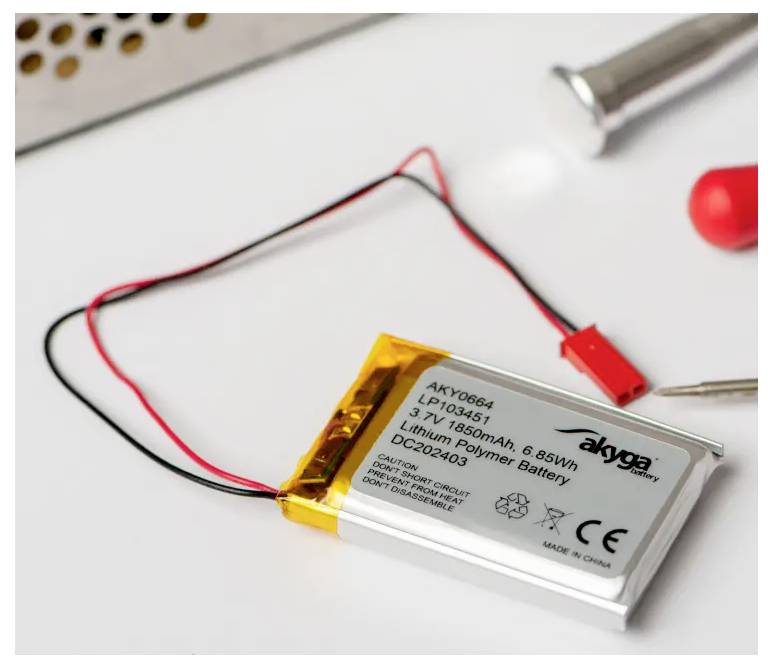 Eine Lithium-Polymer-Batterie mit roten und schwarzen Kabeln liegt auf einem weißen Hintergrund. Links oben ein Teil eines Metallgehäuses.